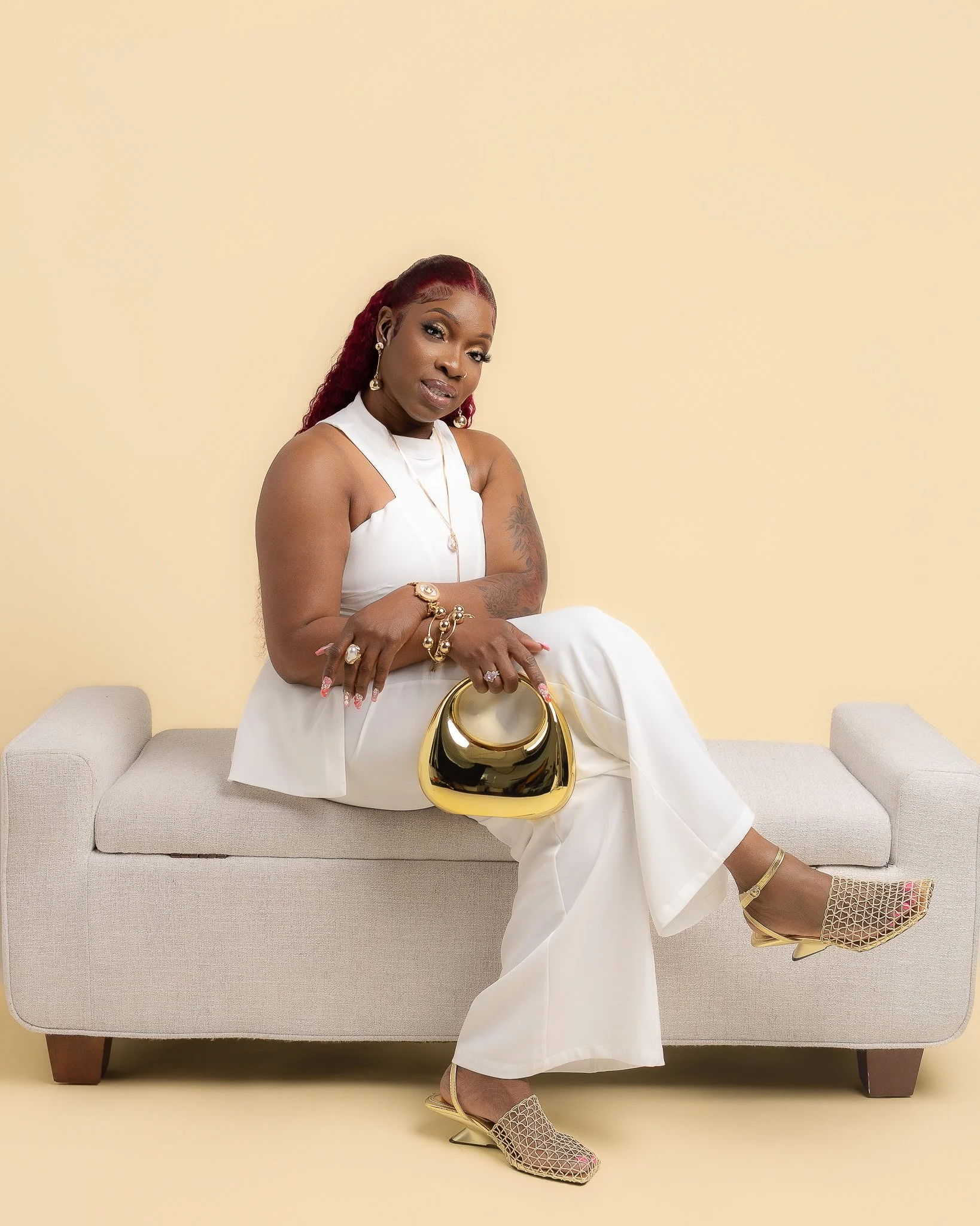 Woman with red hair, wearing white dress, gold jewelry, beige high-heeled shoes, sitting on a beige sofa against a plain light yellow background, holding a gold handbag.