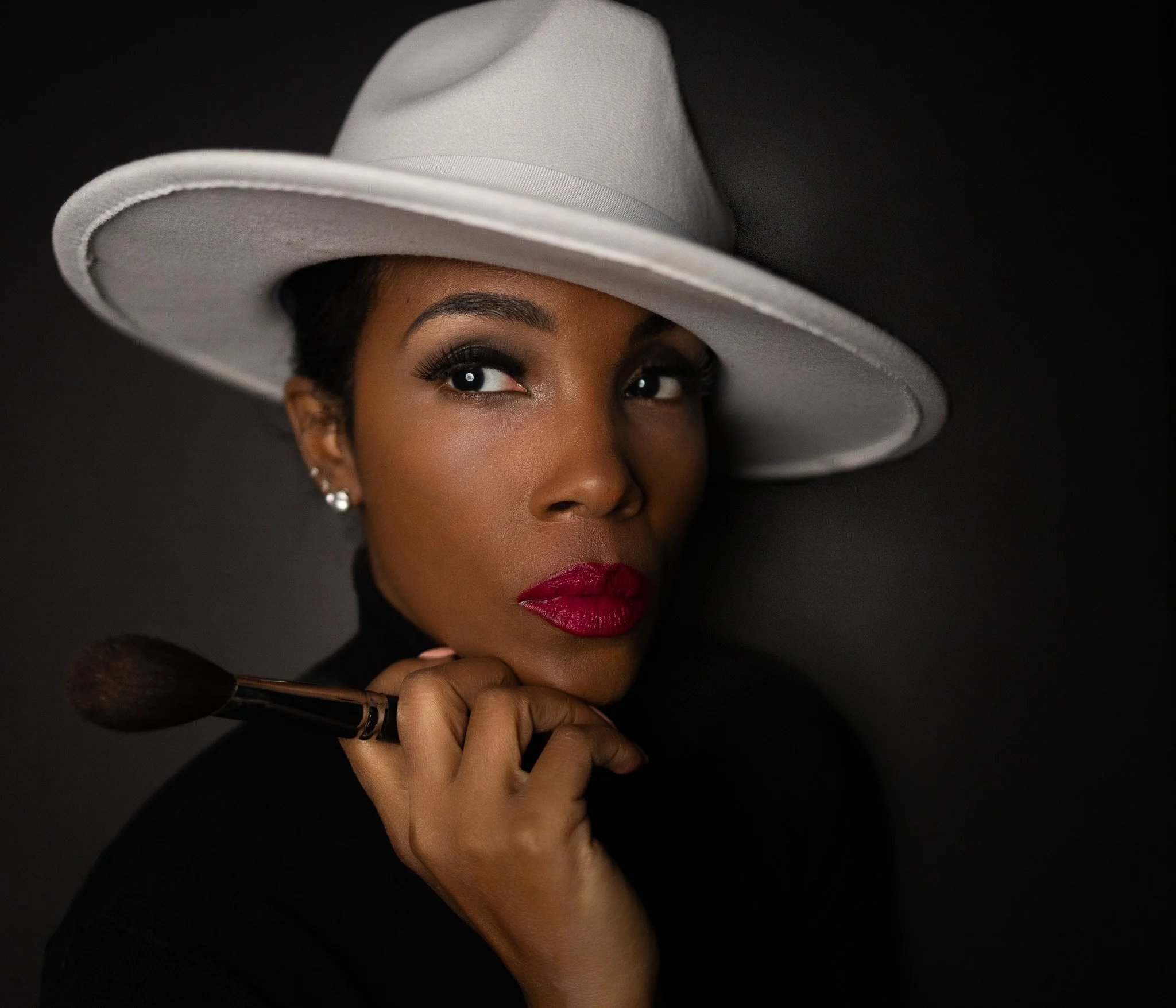 A woman with dark skin and styled eyebrows is wearing a large white hat and red lipstick. She is holding a makeup brush near her chin, and the background is black.