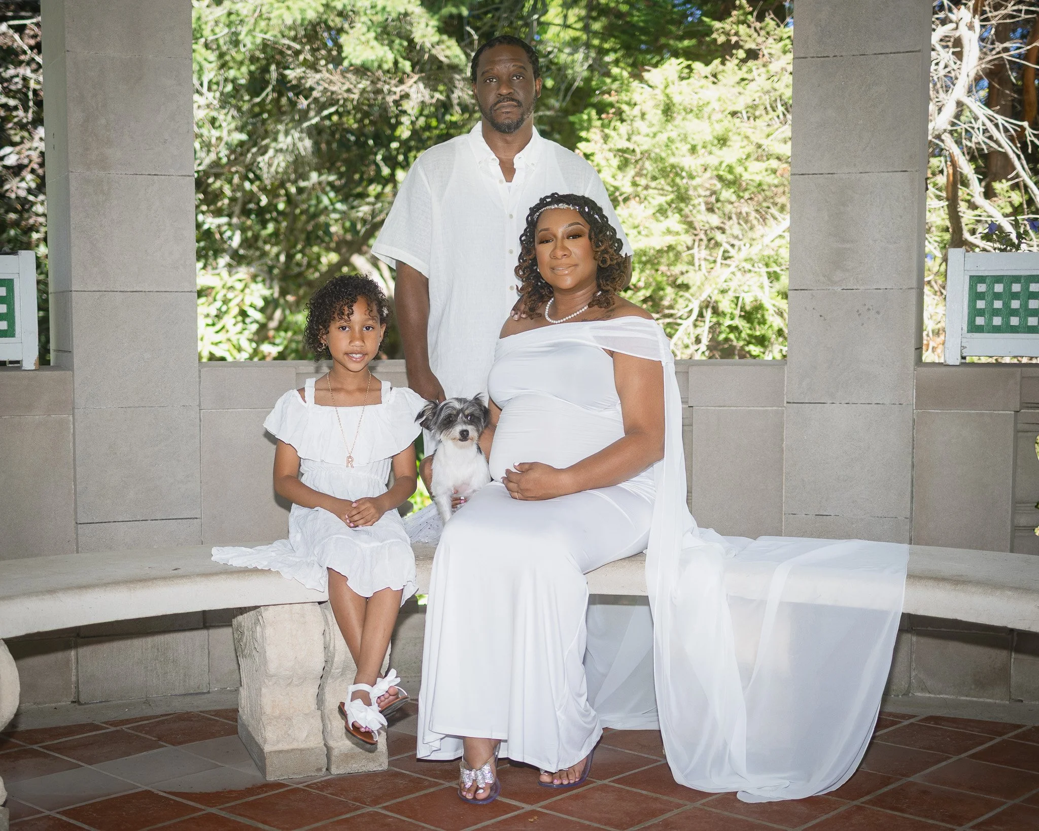 A family of three people and a dog sitting and standing on a stone bench outdoors with leafy trees in the background. The woman is pregnant, wearing a white dress and necklace. The young girl is dressed in a white dress with a letter "R" necklace, an