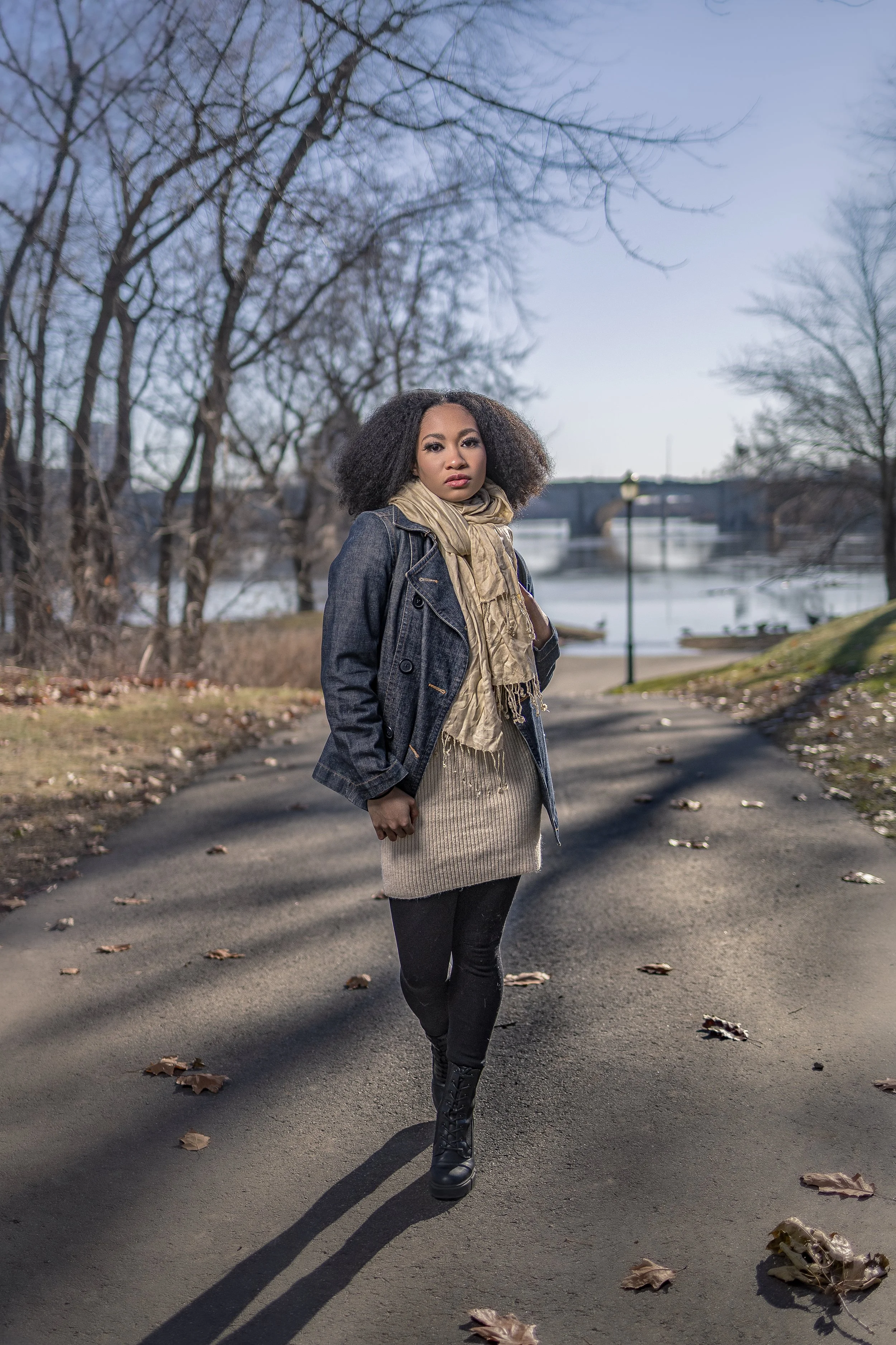 Young woman walking on a paved path in a park during fall, wearing a denim jacket, beige scarf, sweater, black leggings, and boots, with trees and a bridge in the background.