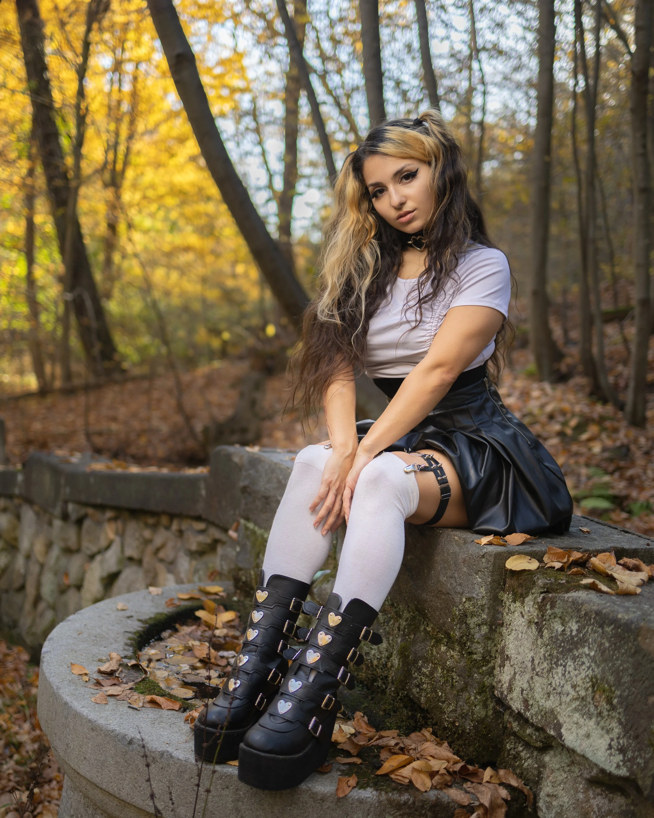 A young woman with dark and blonde wavy hair sitting on a stone bench in a wooded area during fall, wearing a white top, a black leather skirt, white thigh-high stockings, and black knee-high boots with heart-shaped buckles.
