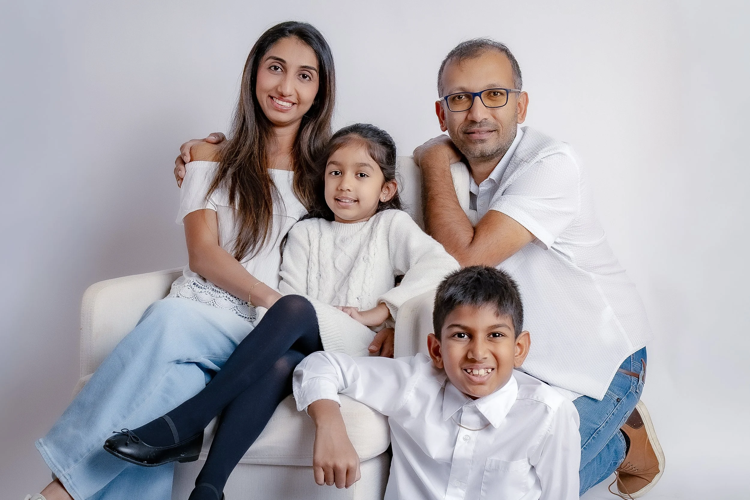 A family of four with two children, a girl and a boy, sitting on a white couch against a plain white wall. The girl, with long dark hair, is sitting on the armrest, smiling, wearing a white sweater and dark tights. The boy, with short dark hair, is s