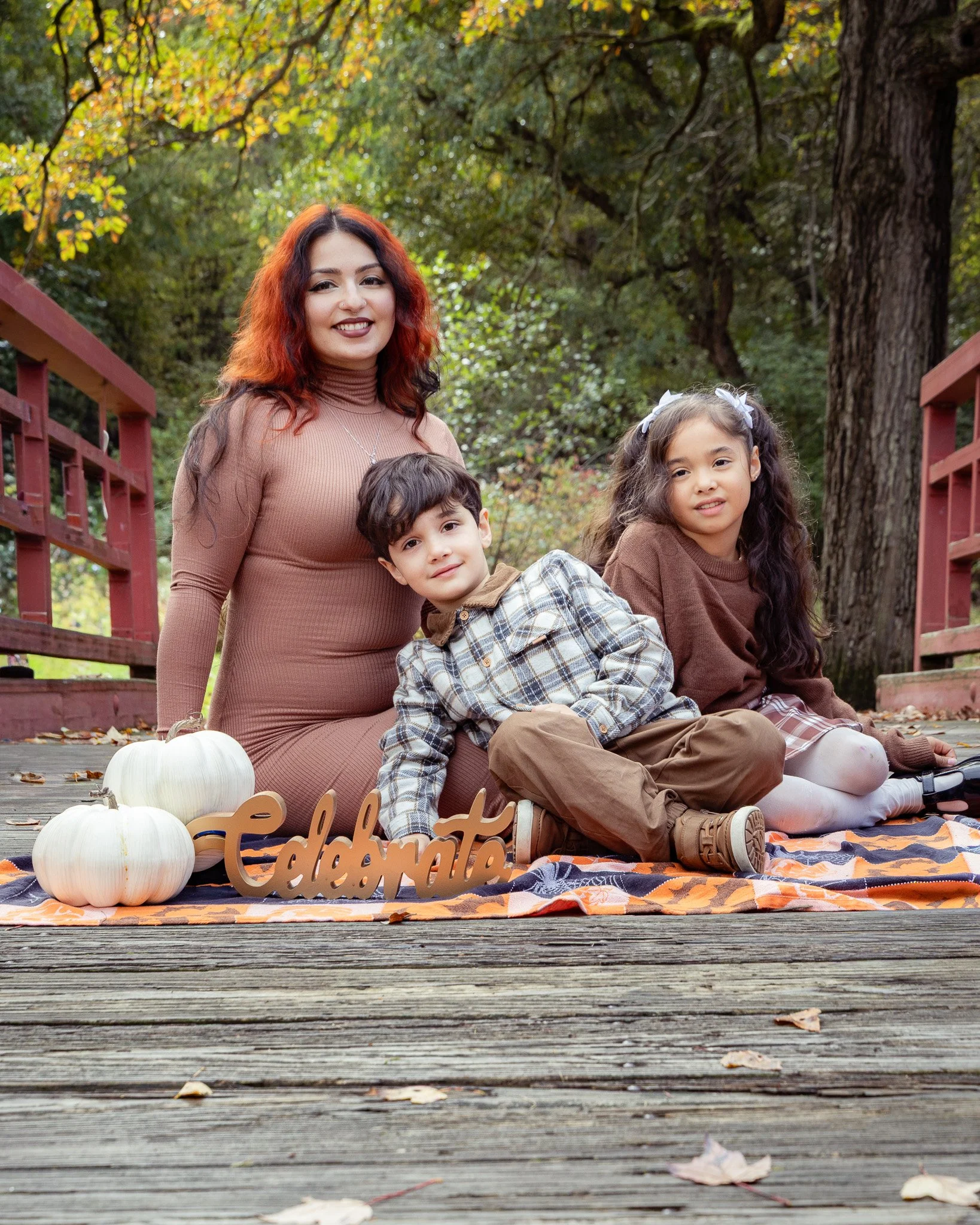 A woman with red hair and two children sitting on a blanket outdoors surrounded by fallen leaves and pumpkins, with a wooded area and large trees in the background, celebrating autumn.