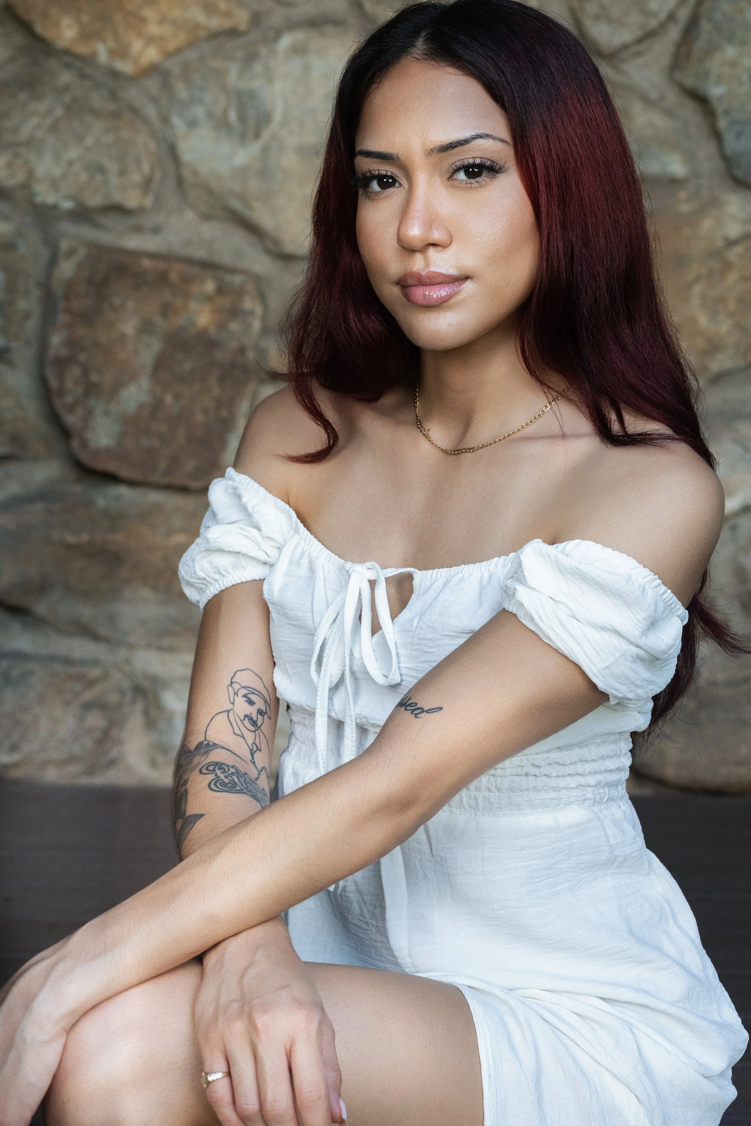 A woman with dark red hair and tattoos on her arms, wearing a white off-the-shoulder dress, sitting with her arms wrapped around her knees in front of a stone wall.