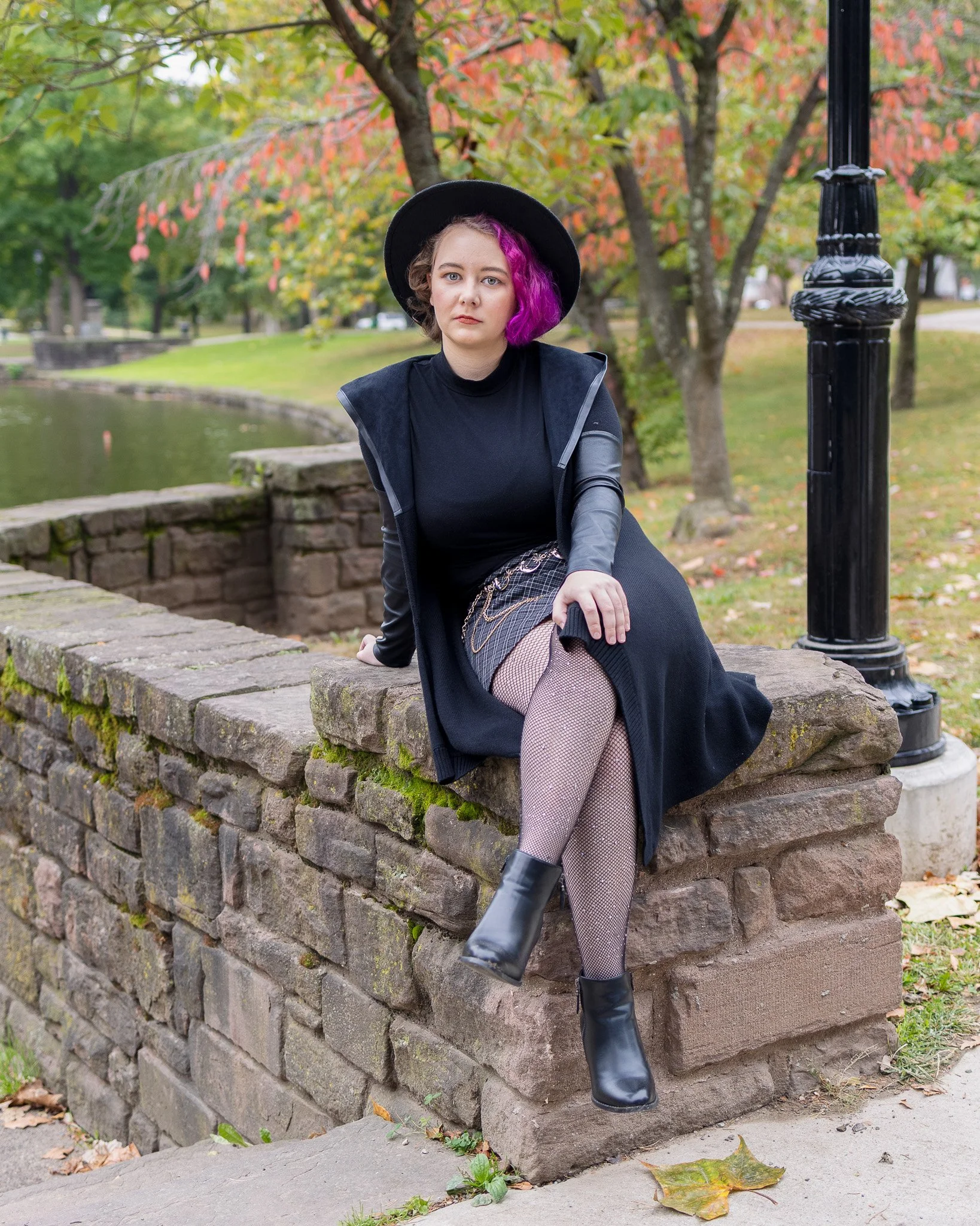 Woman with purple hair, black hat, black jacket, fishnet stockings, and black boots sitting on a stone wall in a park with autumn trees and fallen leaves.