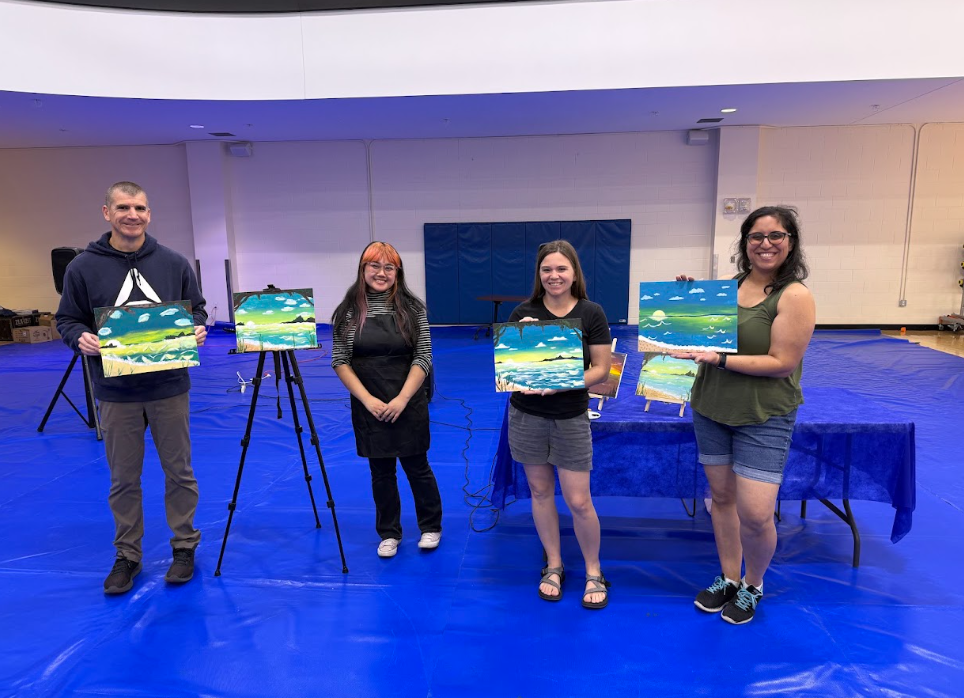 Four people standing in a gymnasium holding and displaying their landscape paintings, with two additional paintings on easels beside them, in front of a blue table and a large blue mat on the floor.