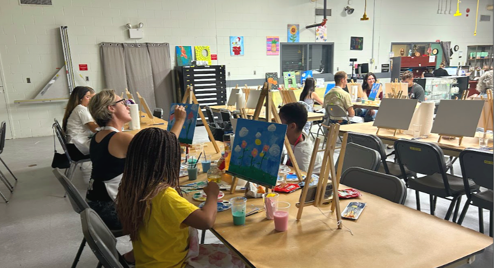 Children and adults painting colorful artwork on canvases in an art classroom.
