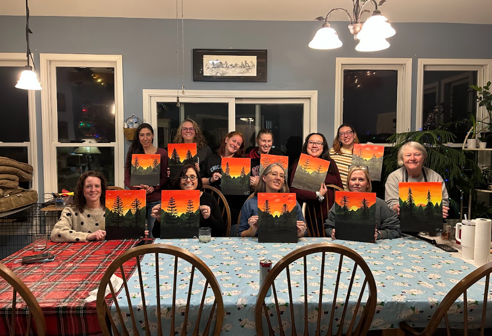 Group of people holding up paintings of a sunset over mountains with trees, sitting and standing around a table in a dining room.