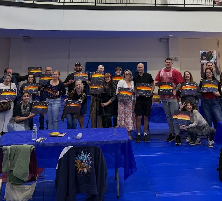 Group of people at an art event holding up paintings of a sunset over mountains, standing behind a table with art supplies, in an indoor hall.