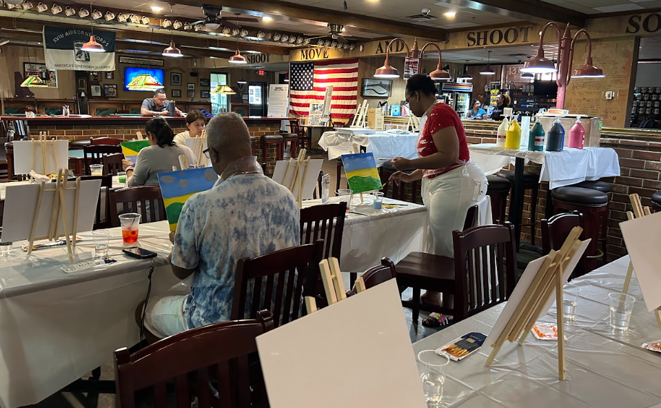 People painting landscapes on canvas at tables in a restaurant-style setting, with an American flag and bar area in the background.