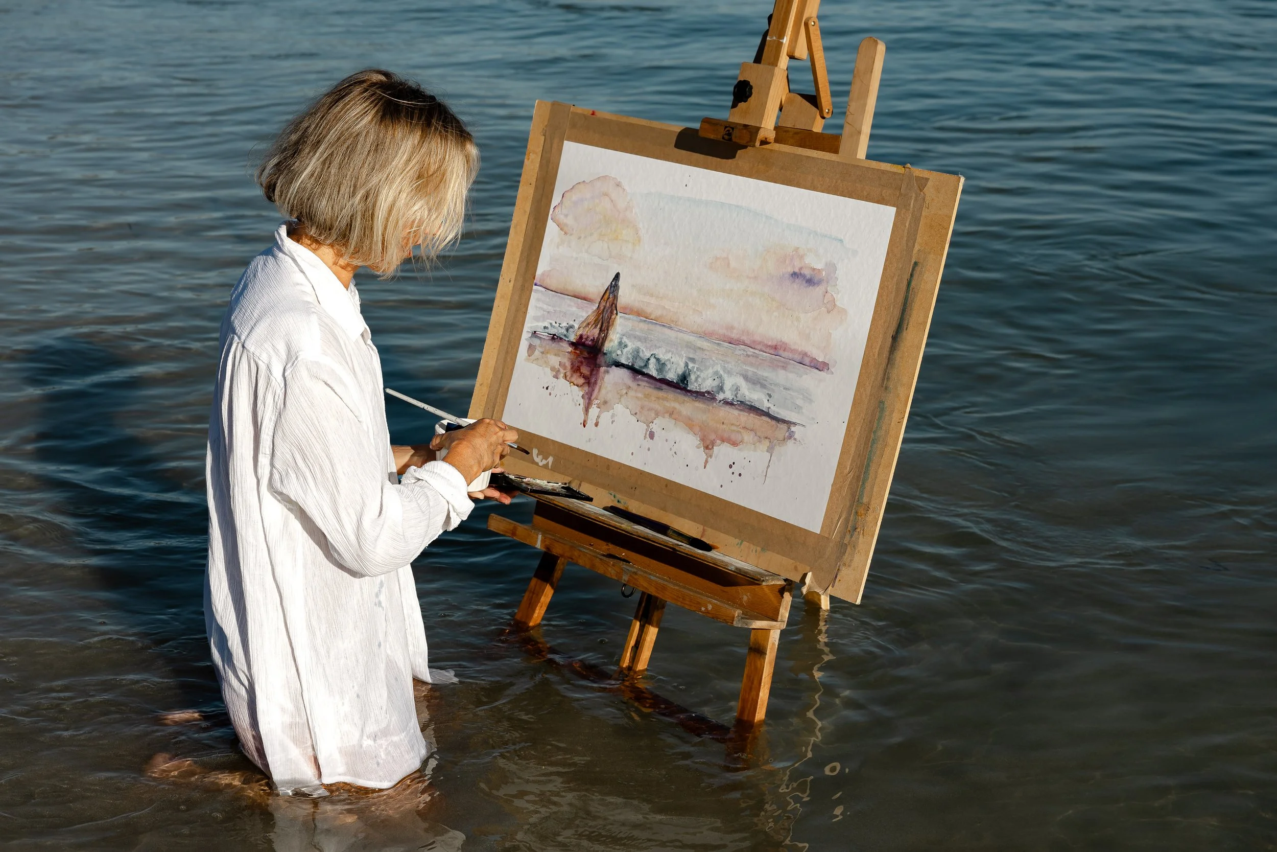 A woman standing in water, painting a landscape on a canvas attached to an easel, with the scene of a sunset over water.