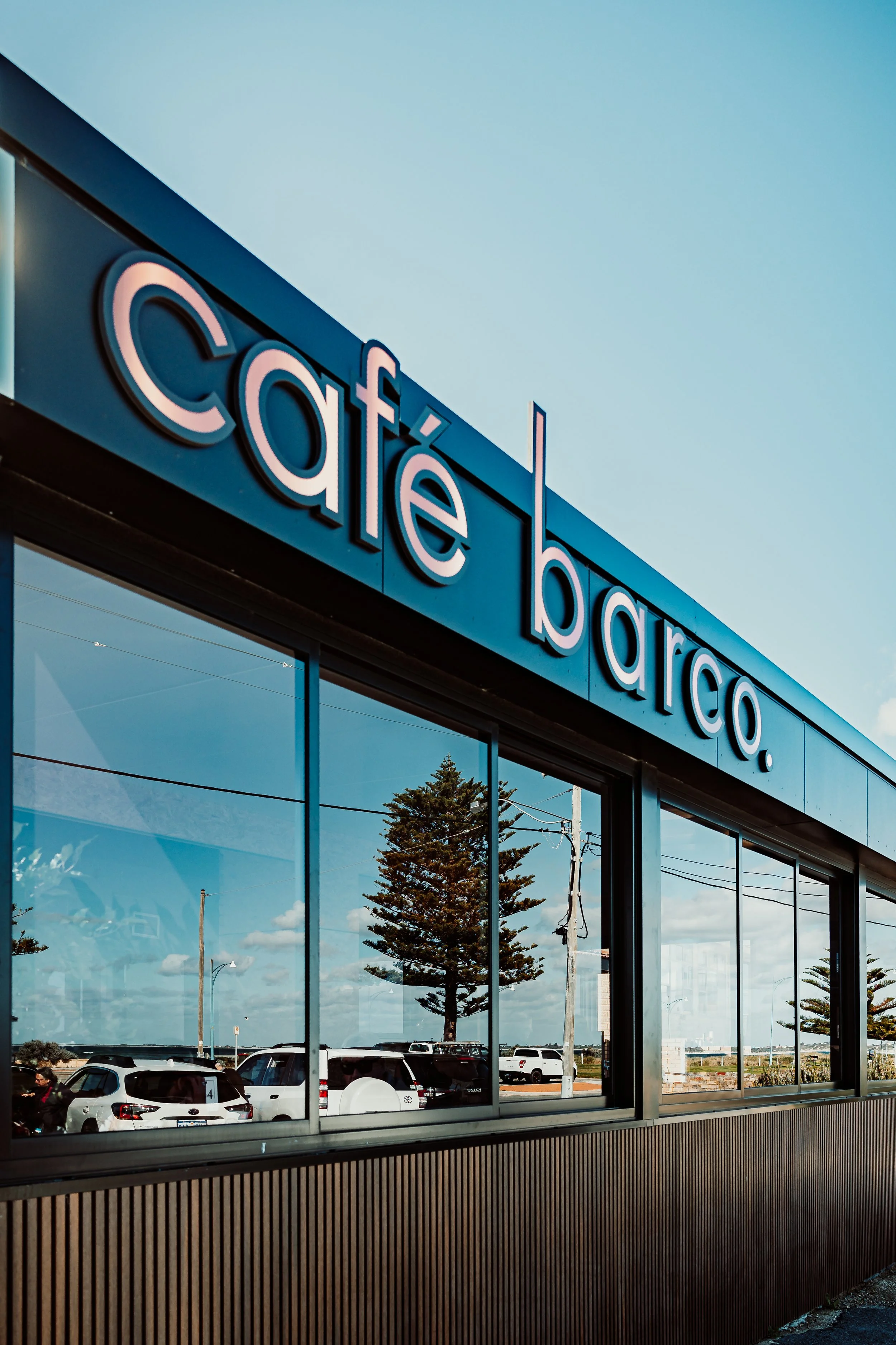 Exterior view of a store with a sign that reads 'Café' in large, illuminated letters, with a parking lot and cars reflected in the glass windows.