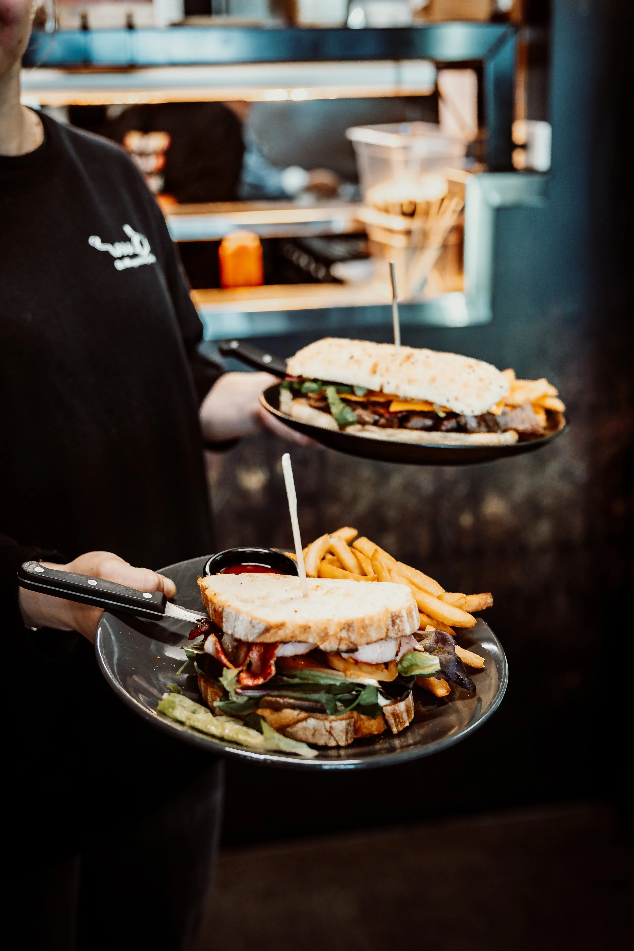 A person holding a tray with a sandwich and fries. The sandwich has various toppings including lettuce, bacon, and sliced bread. The background shows a kitchen or restaurant setting with shelves and containers.