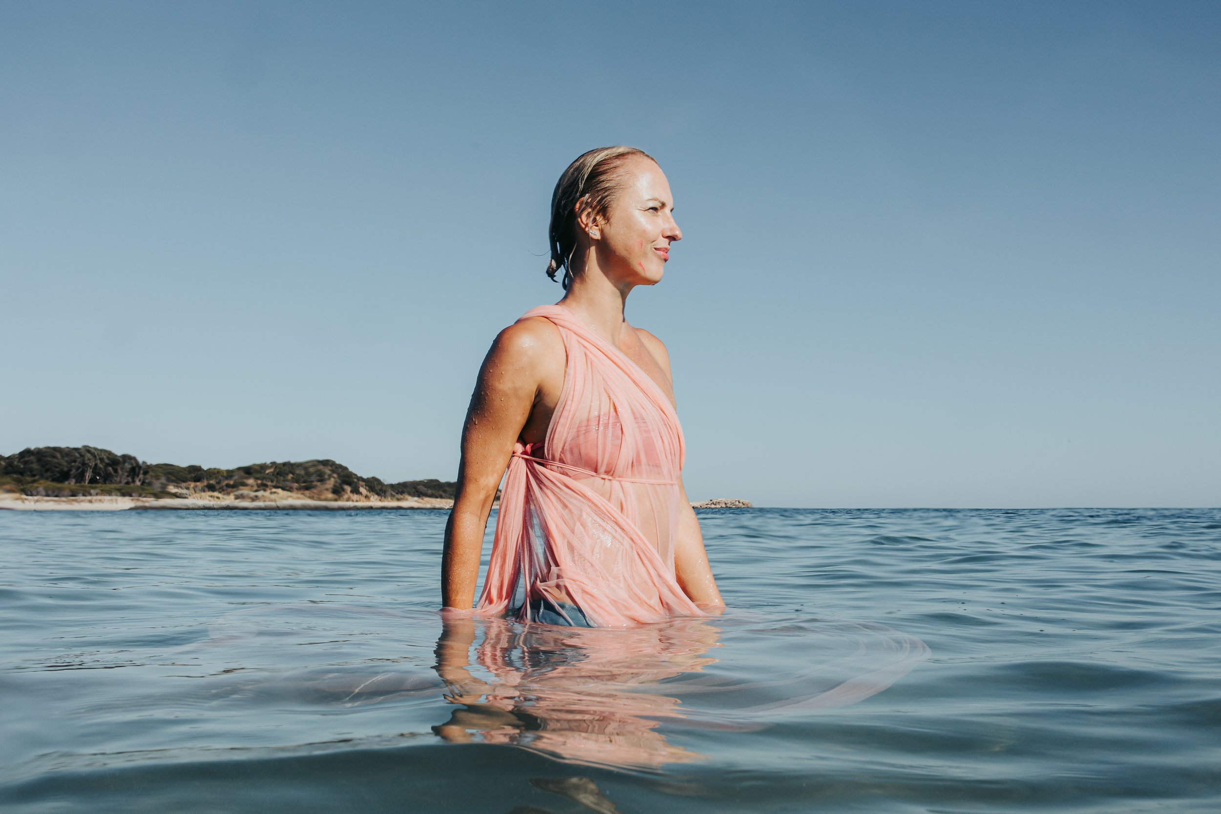 A woman in a pink dress standing in shallow water near a shoreline with trees, under a clear blue sky.