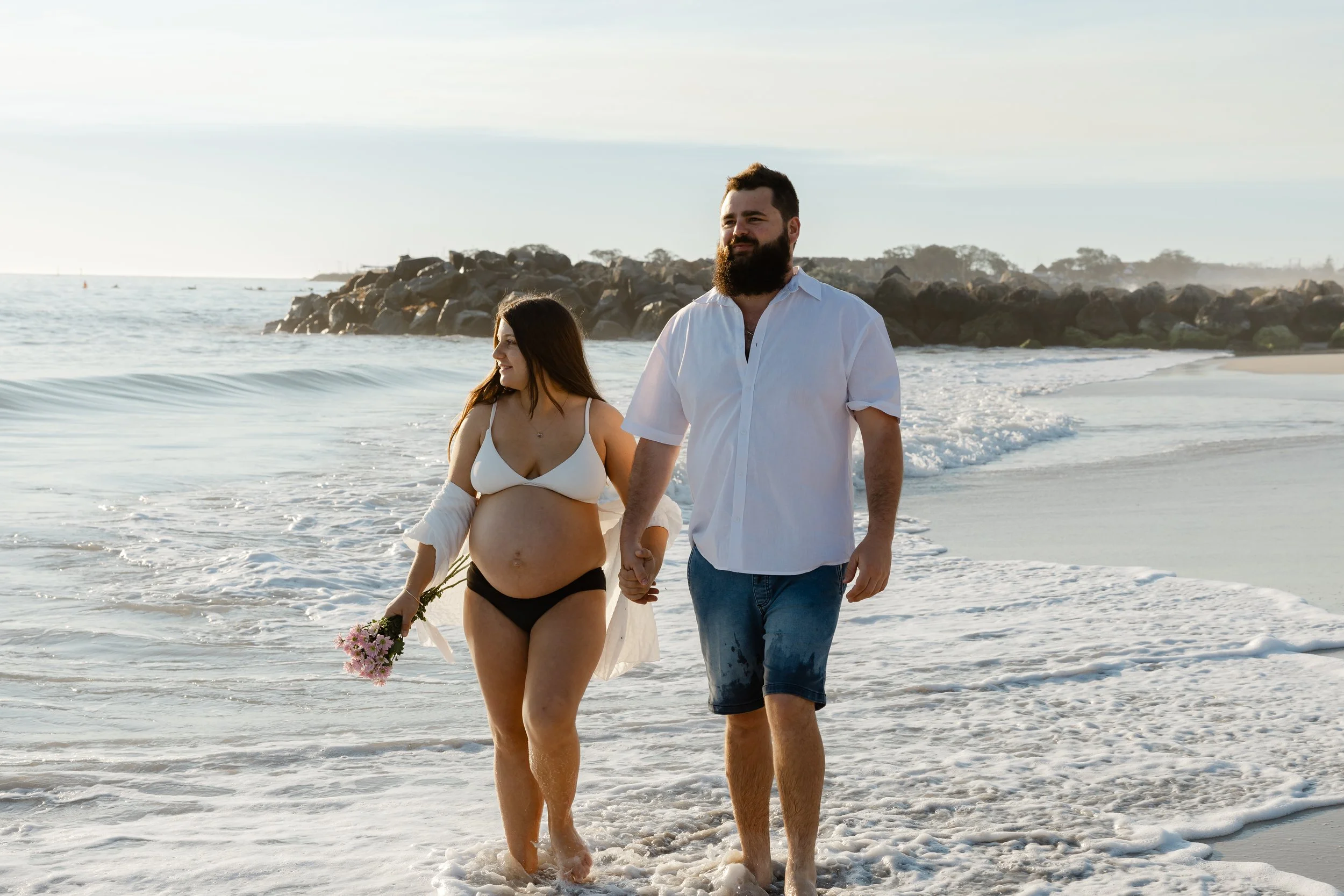 A pregnant woman and a man holding hands walking along the beach at sunset, with waves and rocks in the background.