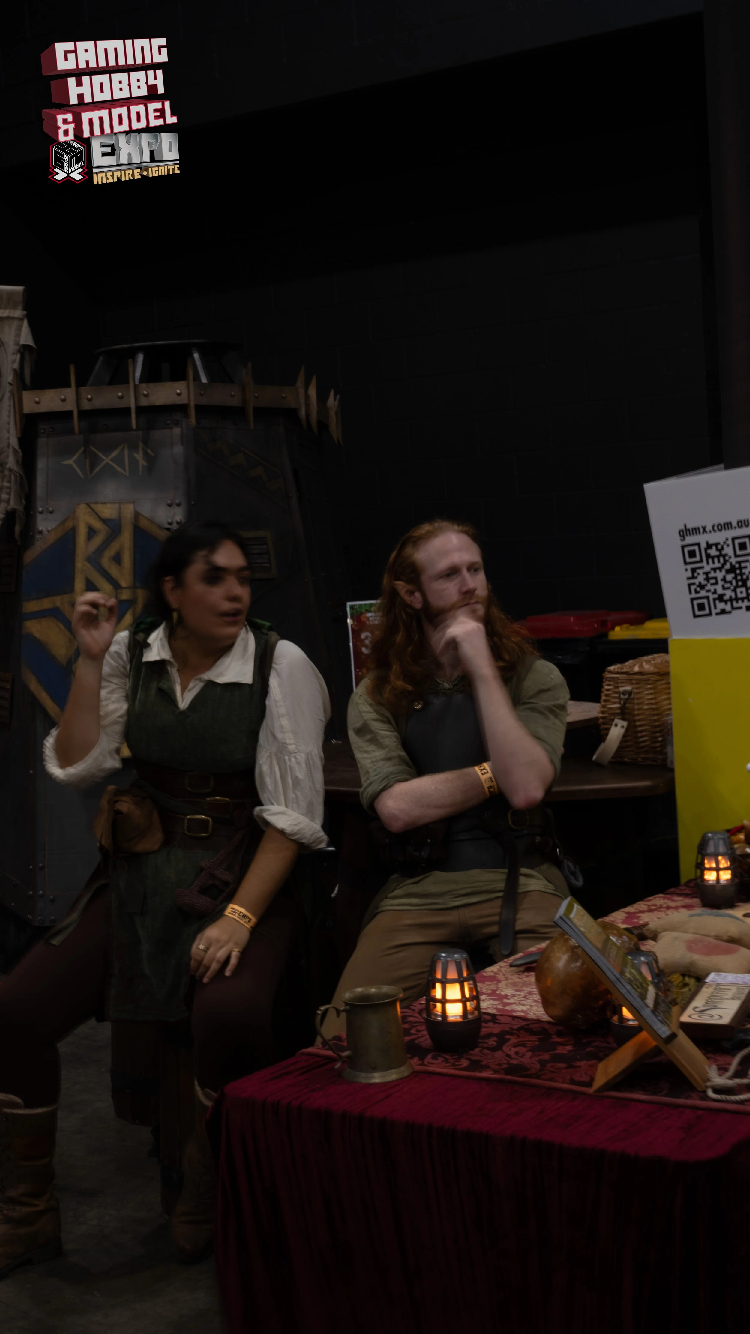 Two people at a booth with medieval or fantasy-themed costumes, sitting behind a table with candles and props, at a gaming and hobby expo.