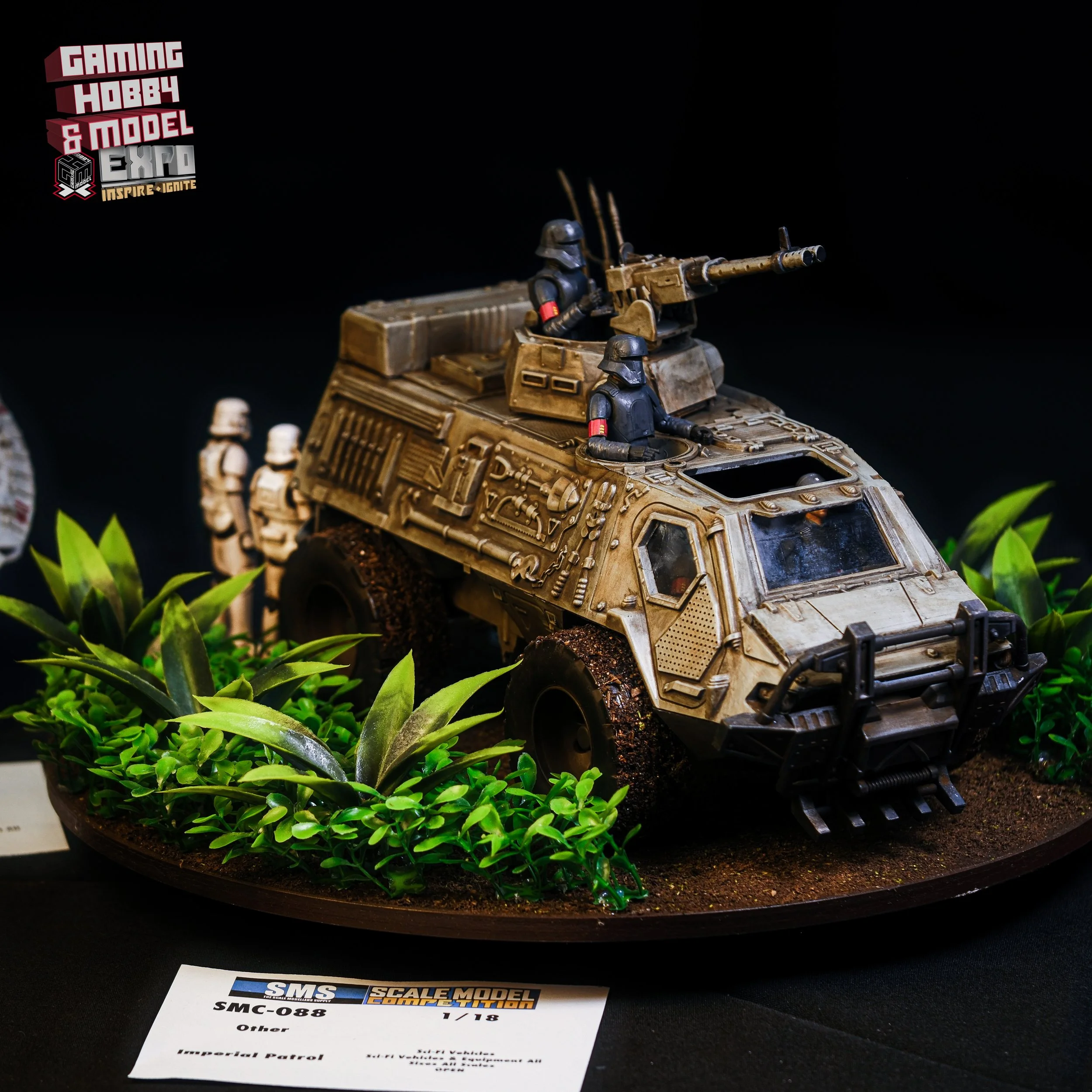 Model of a military armored vehicle with soldiers, displayed on a terrain with green plants, at a hobby and model expo.