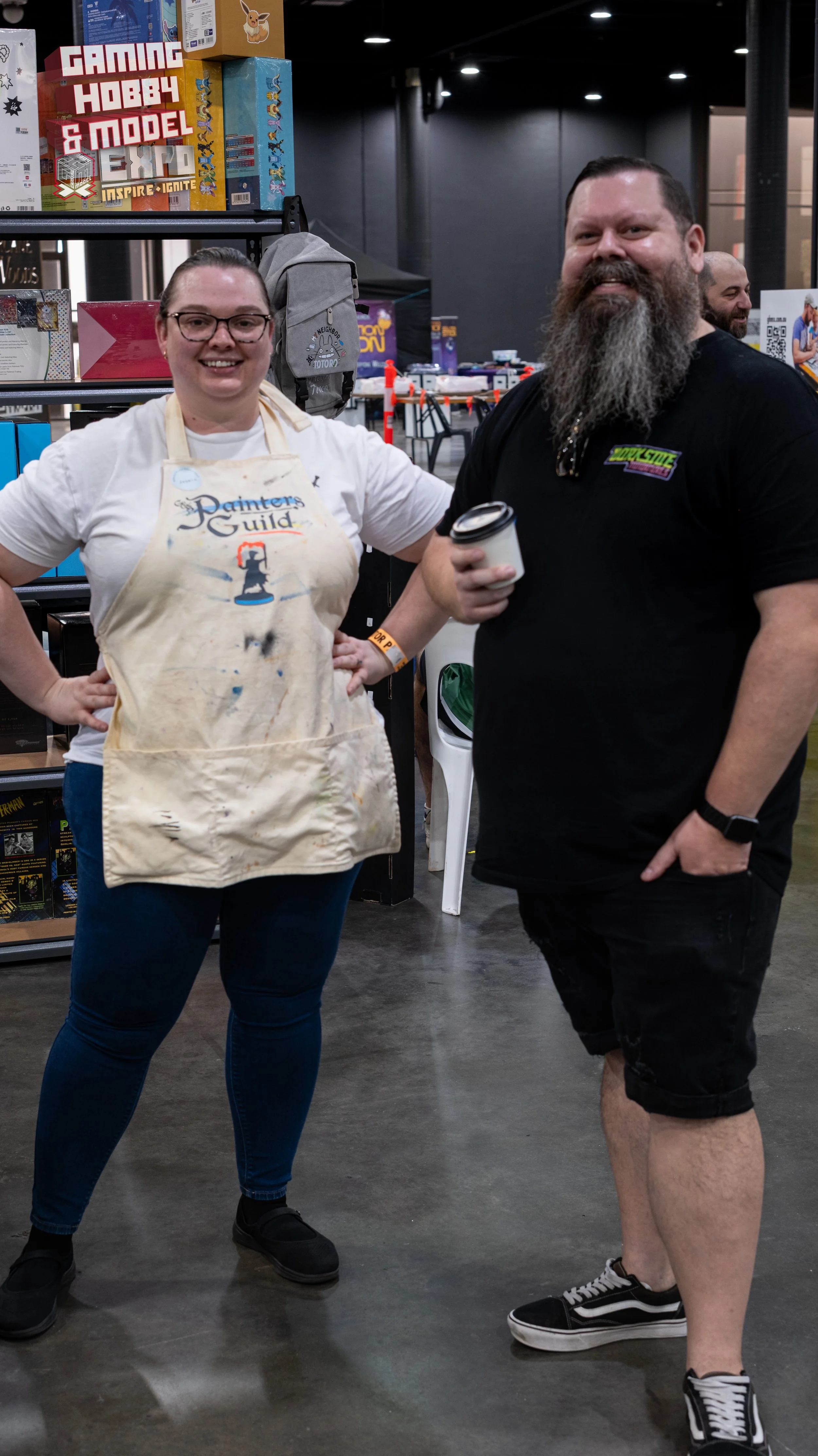 Two people standing inside a store, posing for a photo, with shelves of games and toys in the background, the woman wearing a paint-splattered apron and glasses, and the man with a beard holding a coffee cup.