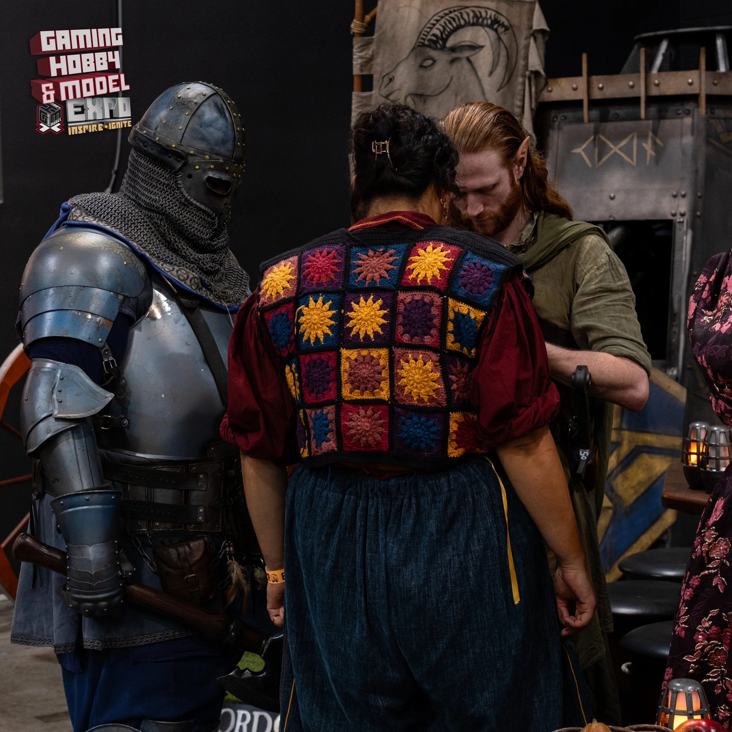 Three people are seen at a convention, with a person dressed in medieval knight armor on the left and a woman wearing a colorful, patchwork crochet vest, and a man with a beard and long hair in conversation.