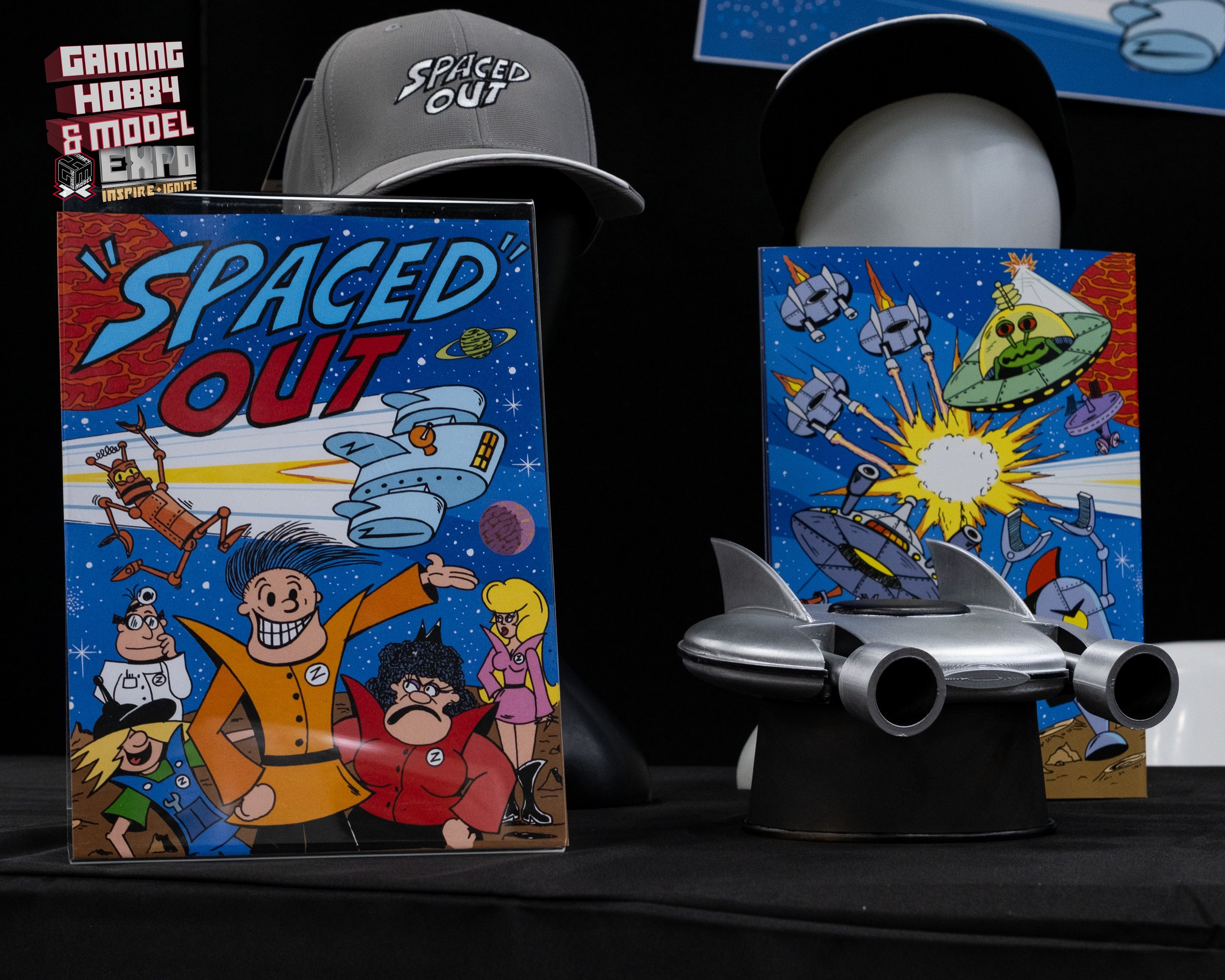 Display of space-themed items including a comic book titled 'Spaced Out' featuring cartoon characters, a gray cap with 'Spaced Out' embroidered, a black hat, a gray spaceship model, and colorful sci-fi artwork in the background.