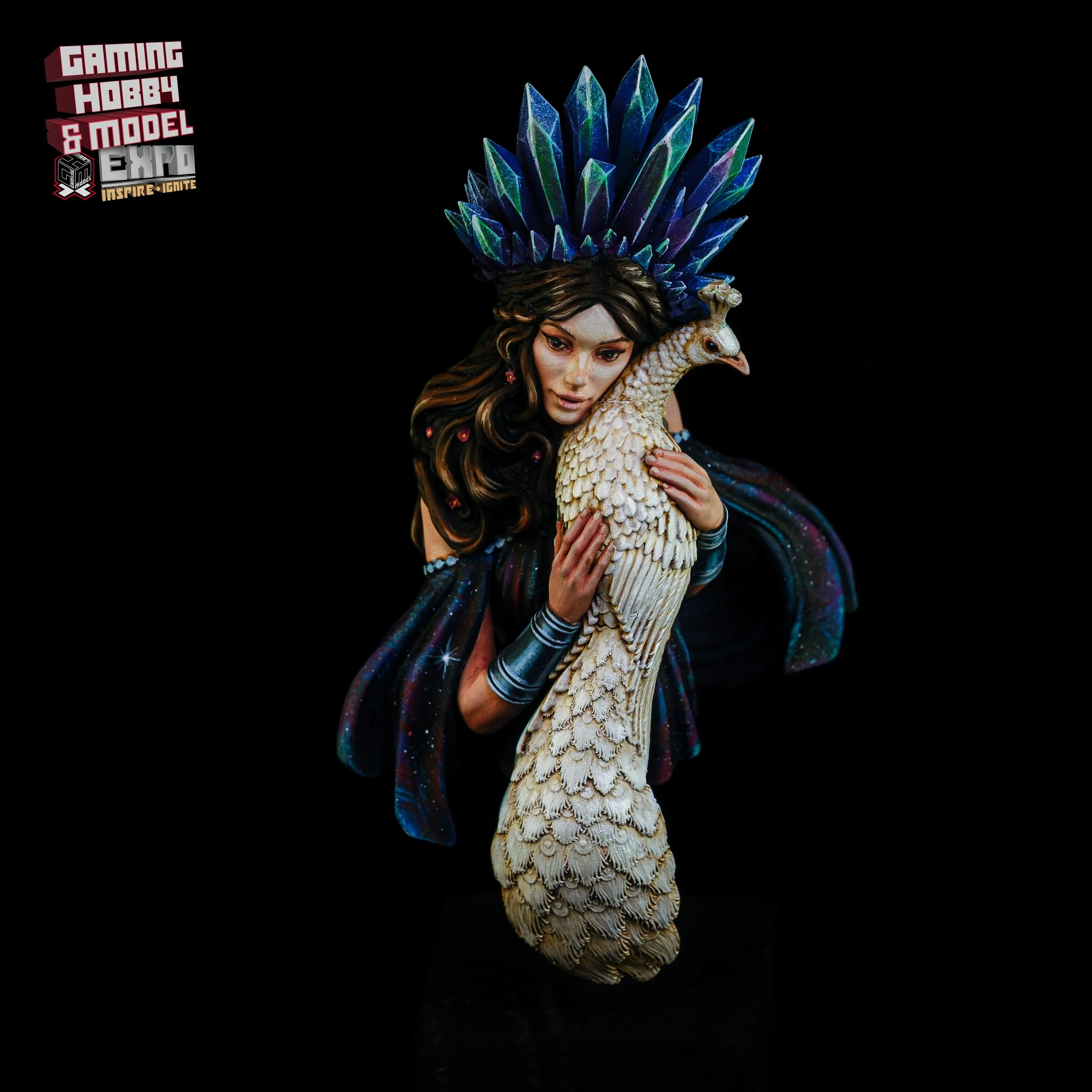 A detailed sculpture of a woman holding a peacock, with a large, colorful headdress resembling feathers, set against a black background, promoting a gaming, hobby, and model expo.