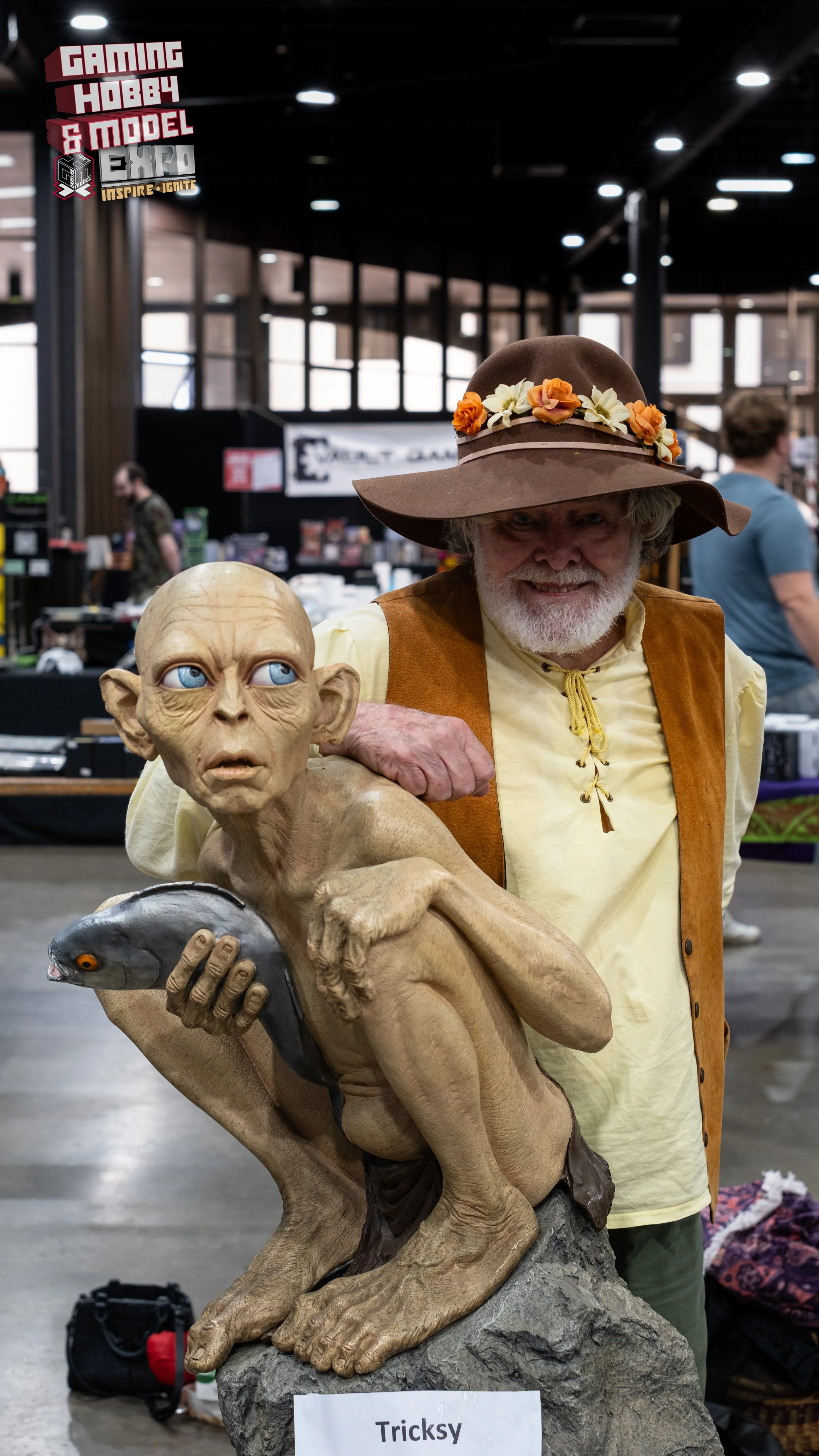 A man dressed as a wizard or fantasy character with a large hat decorated with flowers, standing behind a detailed sculpture of a goblin-like creature with large ears and blue eyes, holding a fish. The sculpture is labeled 'Tricks'. The man is smilin