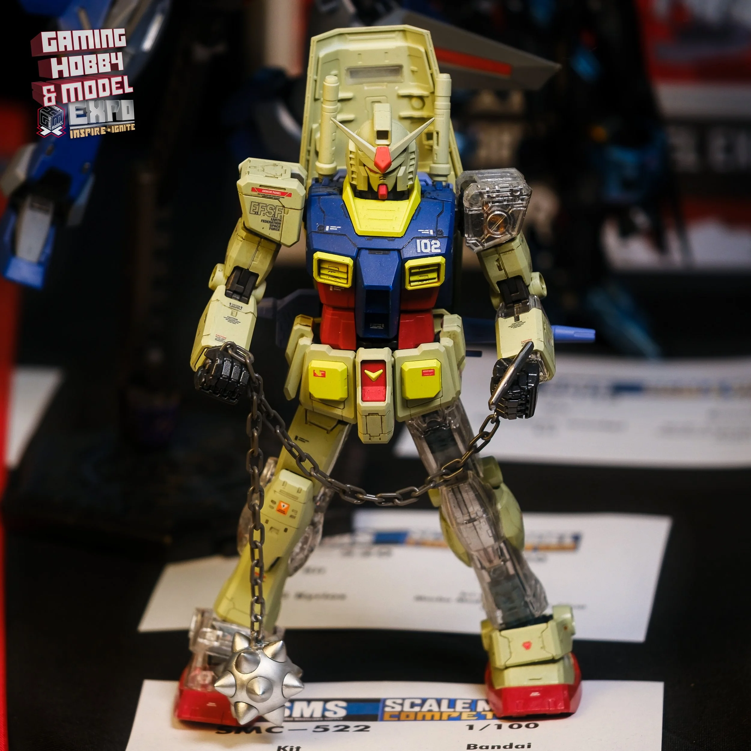 A Gundam model figure standing on a display table, holding a spiked metal ball and chain in its right hand. The model features a yellow, blue, red, and beige color scheme with detailed panel and decal markings.