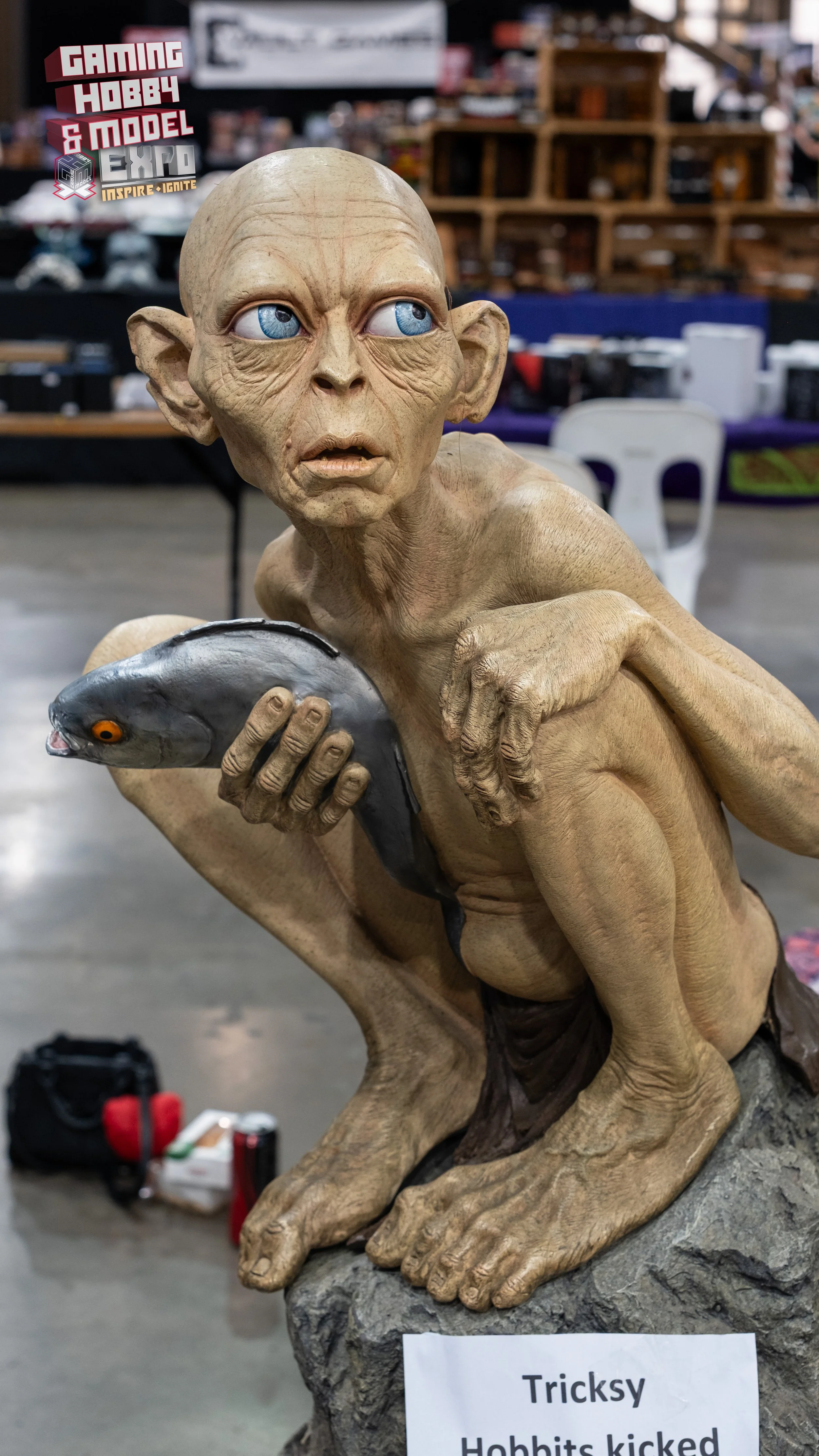 A detailed sculpture of a hobbit holding a fish, displayed at a hobby and model expo. The hobbit has large ears, a wrinkled face, and blue eyes, with a sign in front reading 'Tricksly Hobbits kicked'.