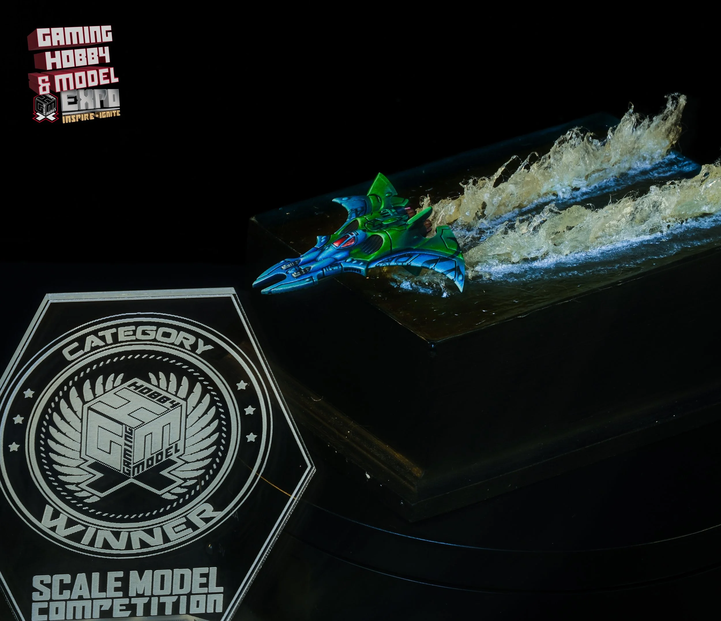 A detailed scale model of a futuristic jet boat racing on water, displayed on a black platform at a competition event, with a 'Category Winner' award plaque in the foreground and a logo in the top left corner reading 'Gaming Hobby & Model Expo'.
