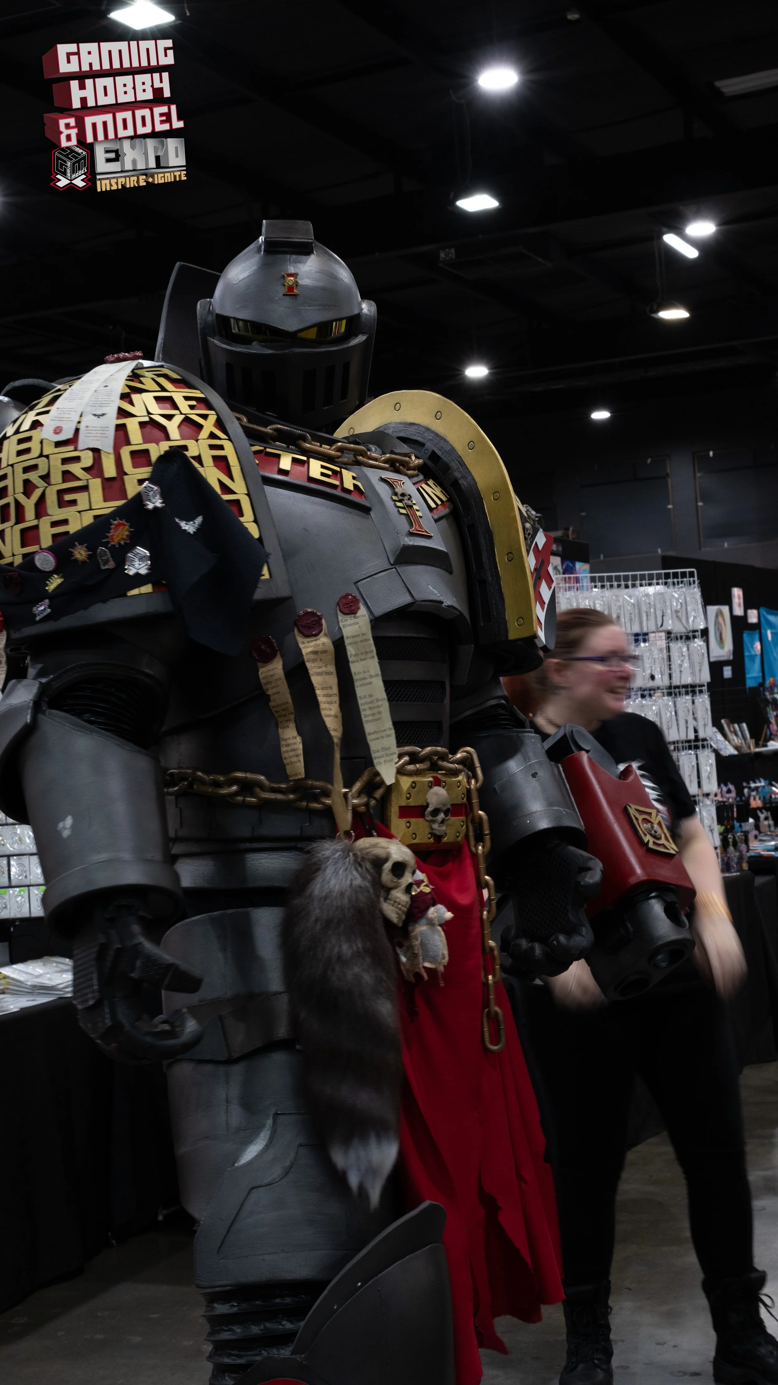 Large armored figure dressed as a Warhammer 40,000 Space Marine at a gaming and model expo, with badges and relics attached. The background shows booths and a woman walking past.