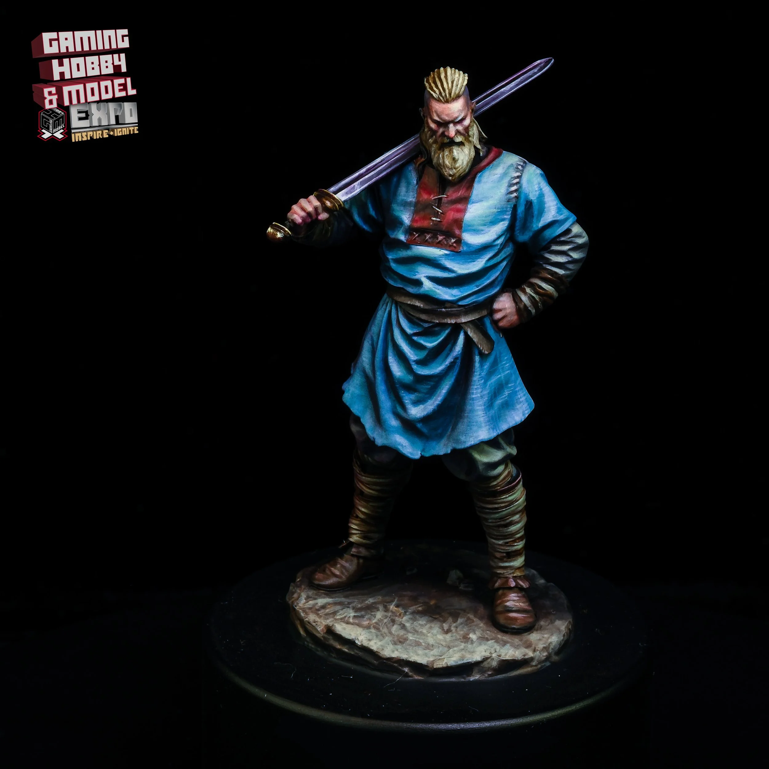 A detailed miniature figurine of a medieval warrior with a beard, wearing a blue tunic, brown boots, and holding a sword over his shoulder, standing on a rocky base against a black background.