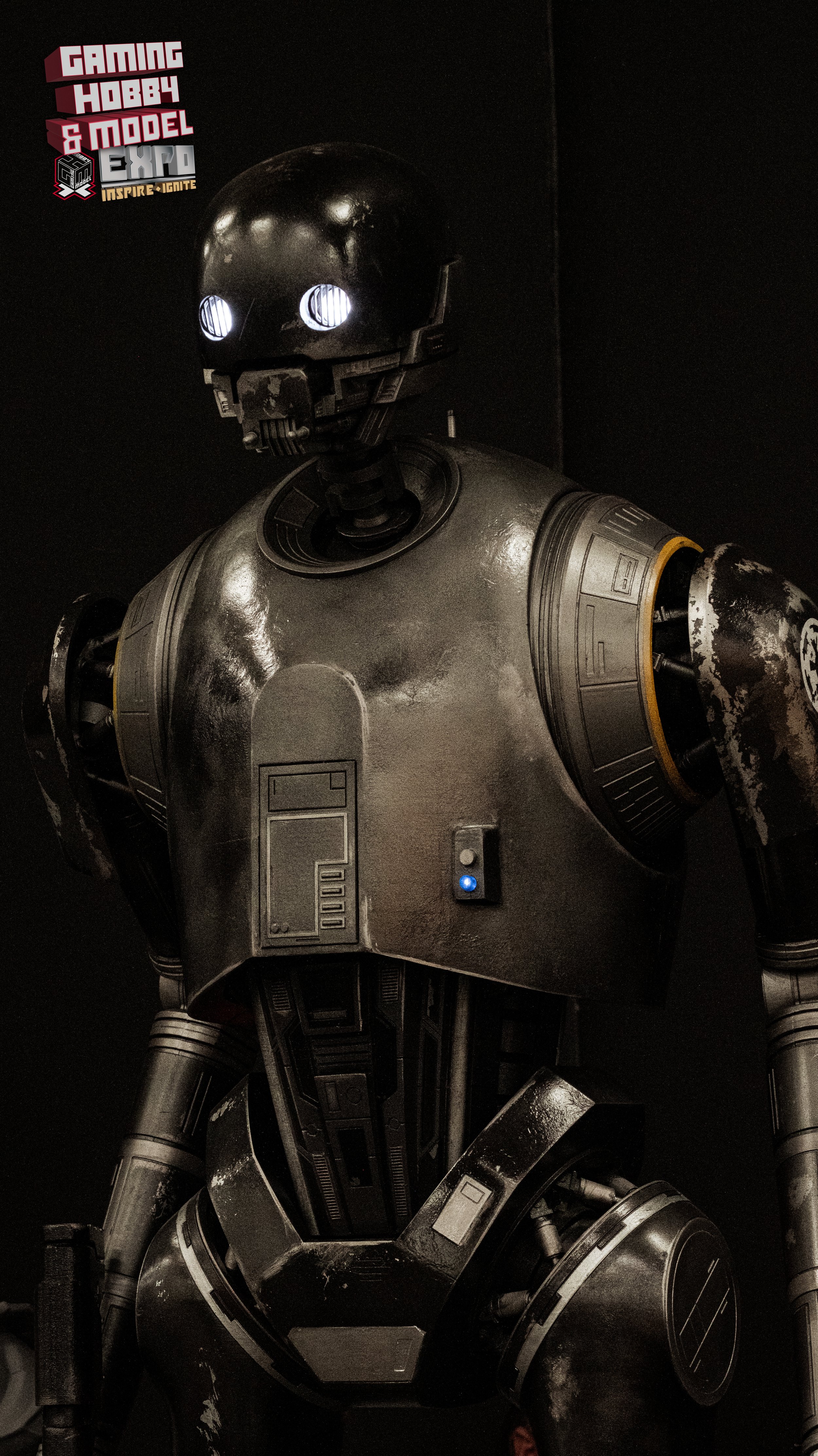 A life-sized replica of the ED-209 robot from the movie "Robocop" on display at a gaming, hobby, and model expo with a badge and logo on the top left corner.