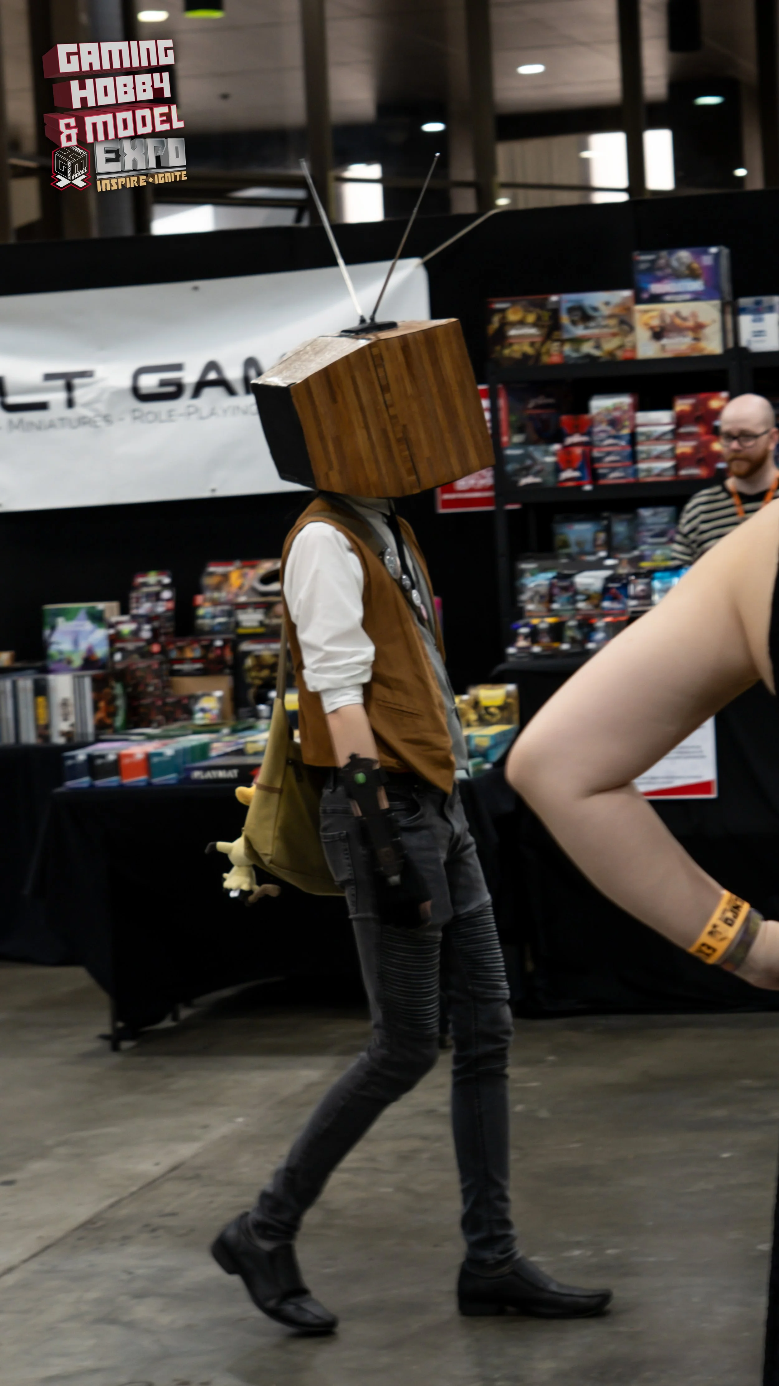 Person walking at a convention wearing a costume with a vintage television set as a head, featuring antennae. The background shows booths with gaming and model merchandise, and there is a banner that reads 'GAMING HOBBY & MODEL'.