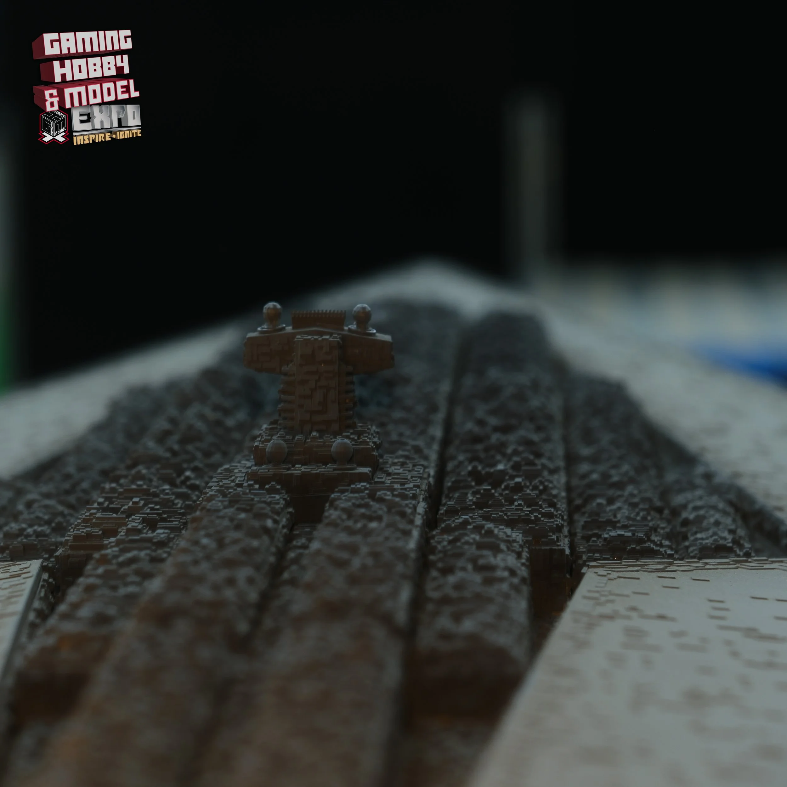 Close-up of a detailed model train set on tracks, with a scoreboard in the top left corner displaying text about a gaming hobby and model expo.