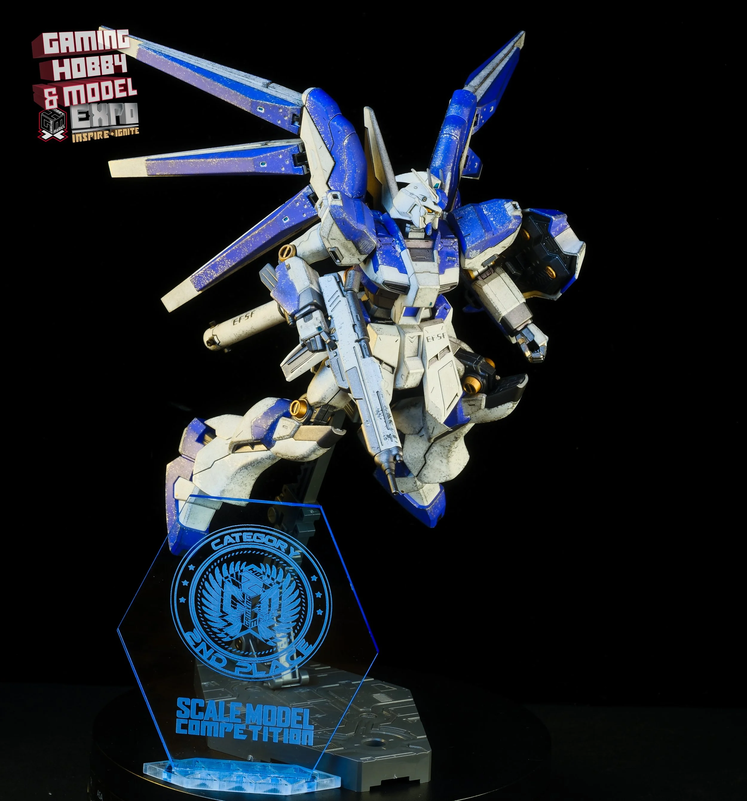 A detailed blue and white Gundam model on a display stand with 'Scale Model Competition' and 'Category 4' labels, set against a black background.