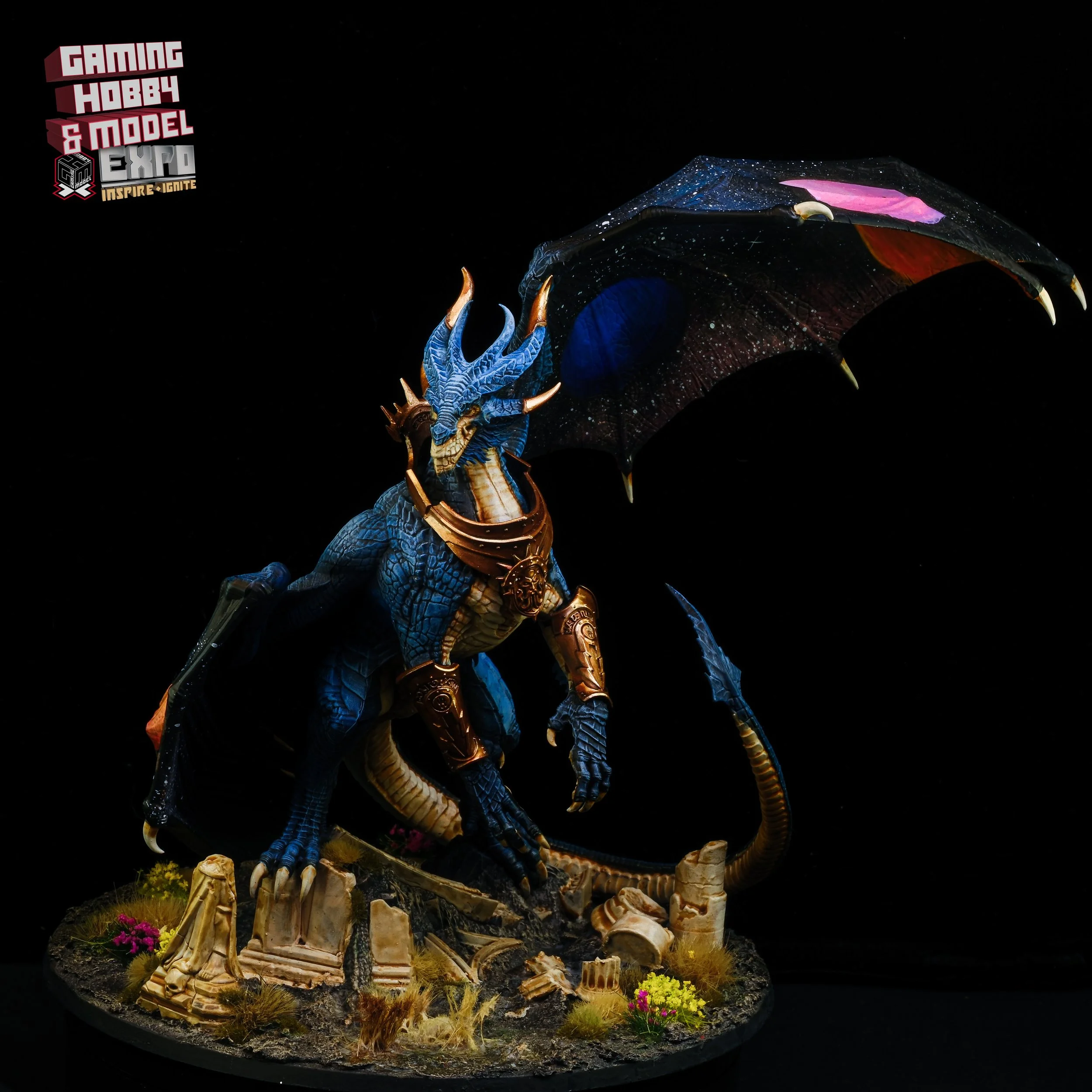 A detailed dragon miniature figurine with blue scales, gold armor, and black wings with starry patterns, standing on a base with ruins and plants, set against a black background, with a 'Gaming Hobby & Model Expo' logo in the top left corner.