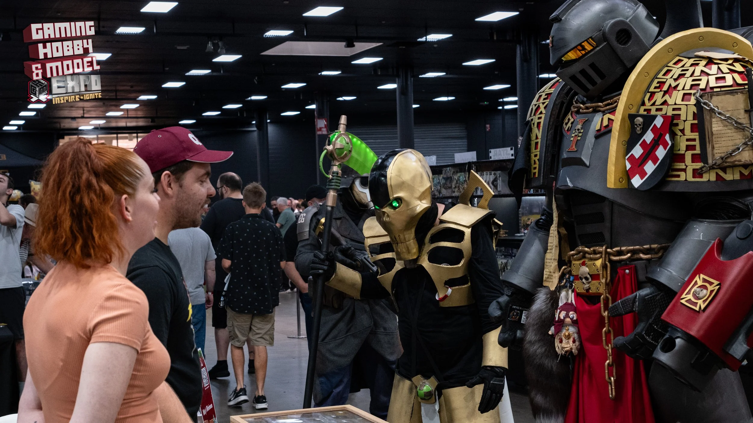 Three people look at individuals dressed as Overwatch characters Reaper and Reinhardt at a convention.