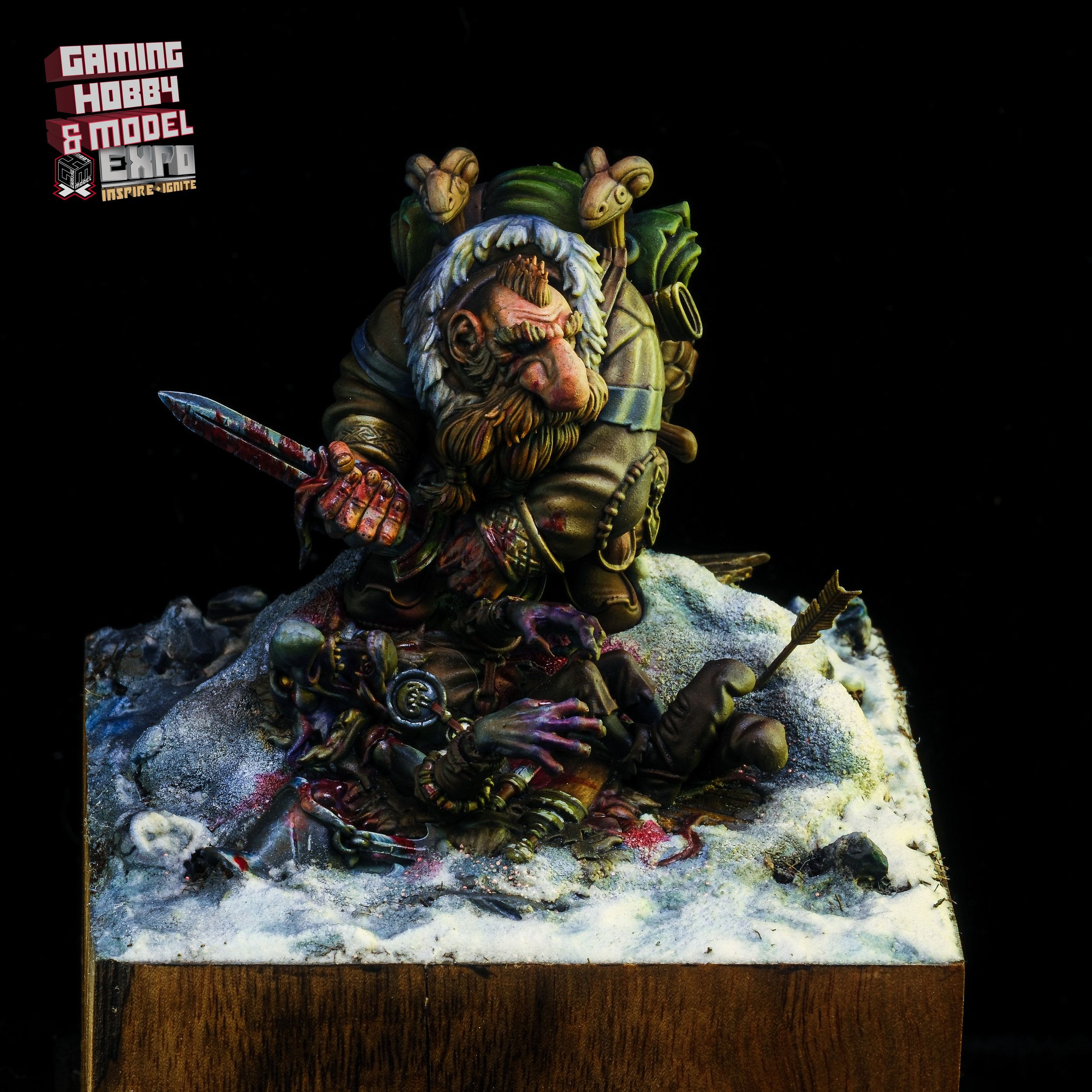 A detailed miniature model of a dwarf with a beard, carrying a backpack and holding a bloodied dagger, standing over a fallen orc with an arrow in its eye, set on a snowy base.