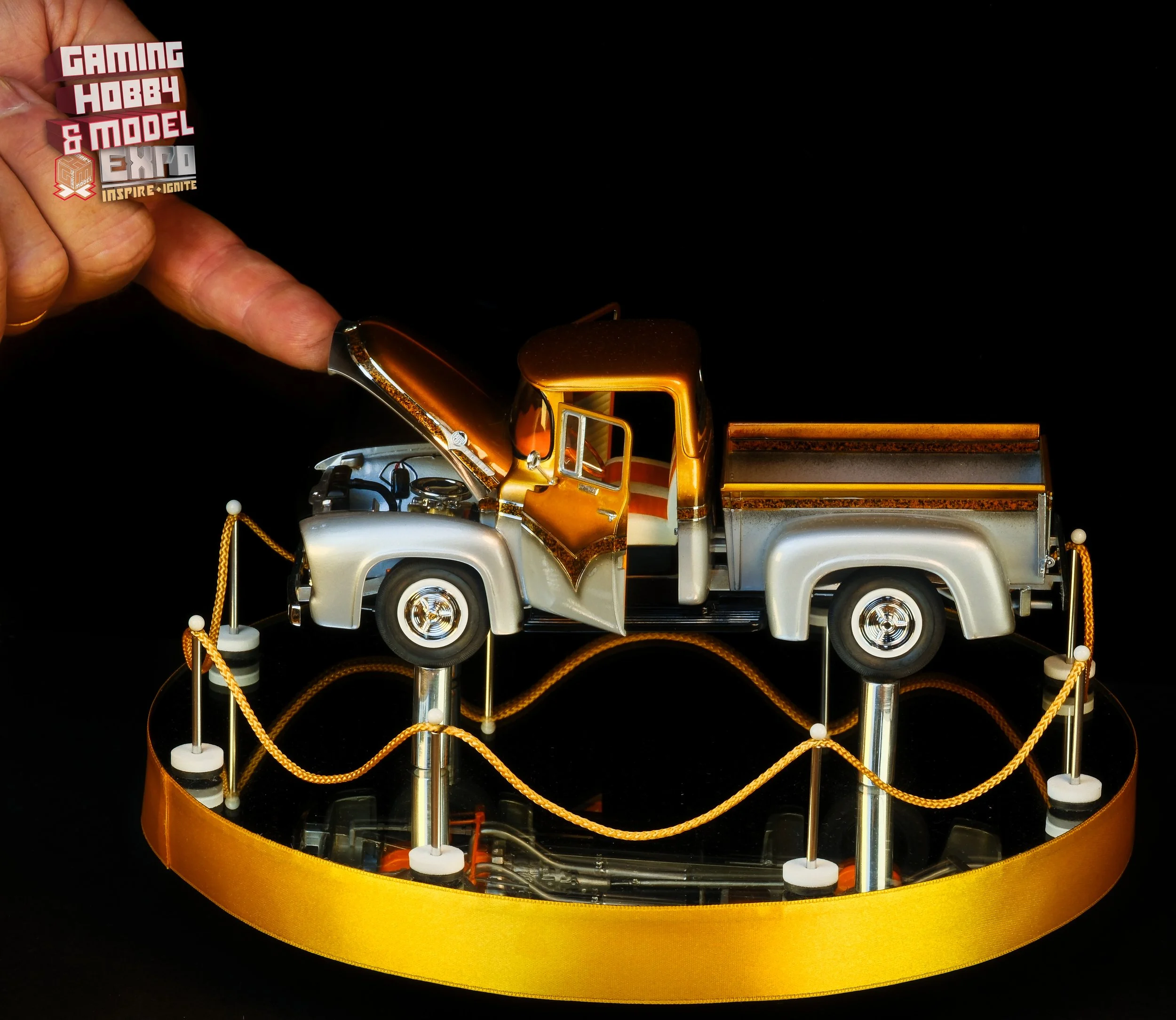 A detailed model of a vintage pickup truck with a silver body, orange accents, and an open hood, displayed on a black reflective surface and surrounded by a yellow ribbon stand.