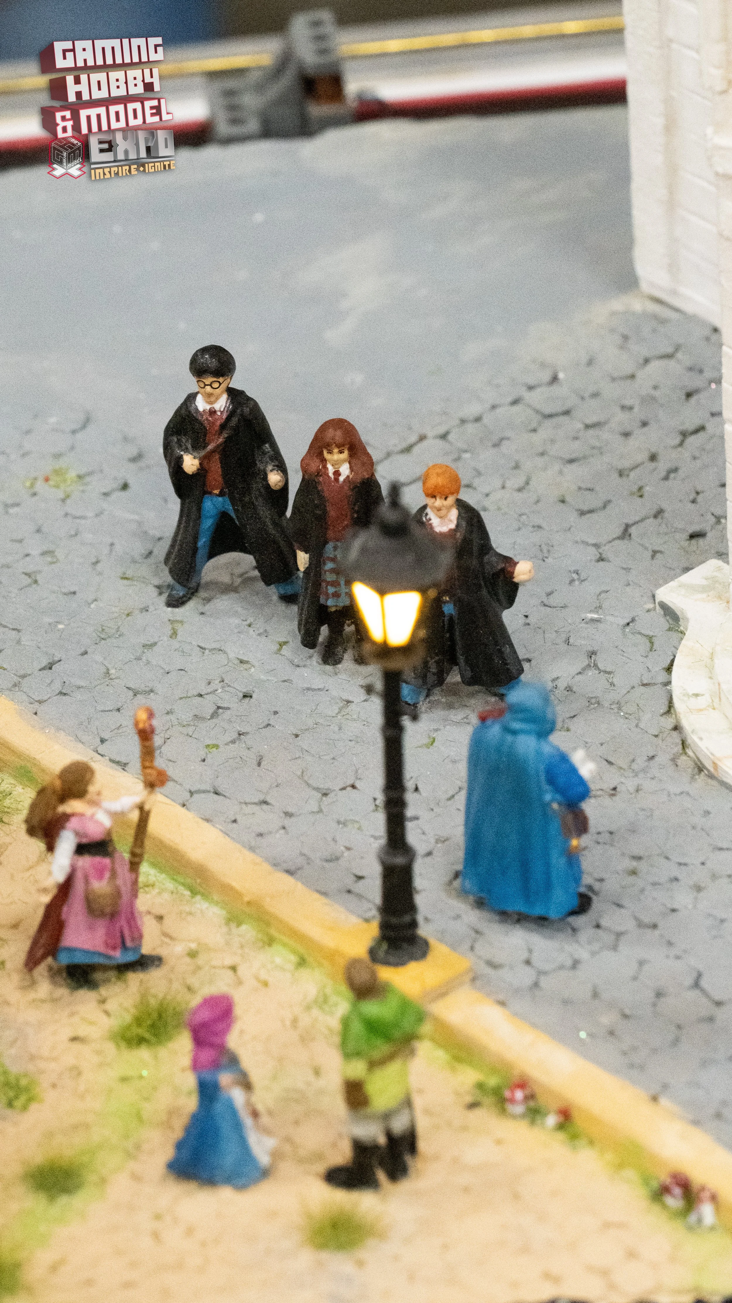 Miniature figures of Harry Potter characters walking on a cobblestone street with a lamp post, near a grassy area with people, a woman in a pink dress, a child in a blue dress, and a person in a high-visibility vest.