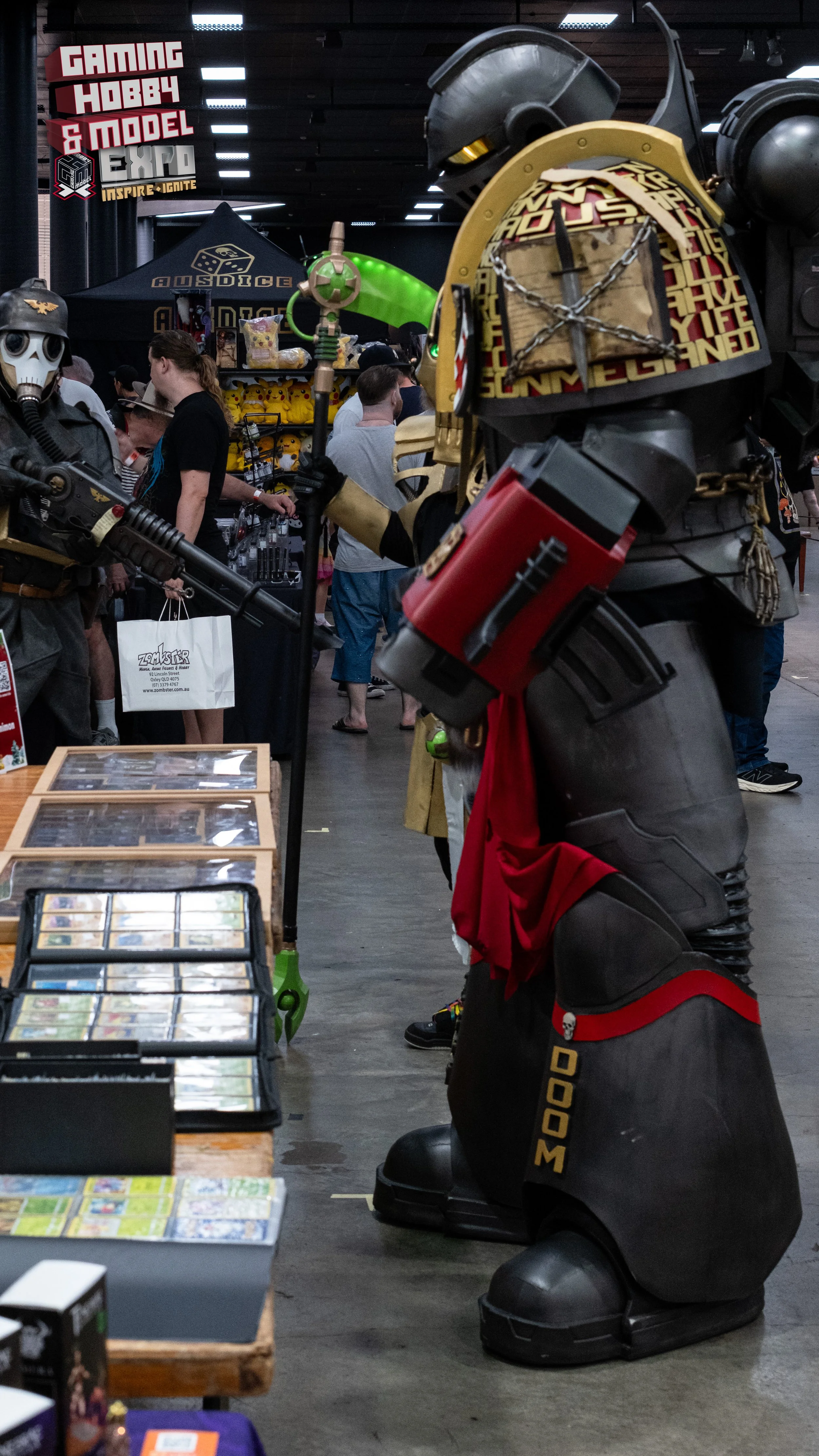 Costumed individuals dressed as characters from the game Overwatch, including Reaper and Bastion, at a convention with booths and attendees in the background.