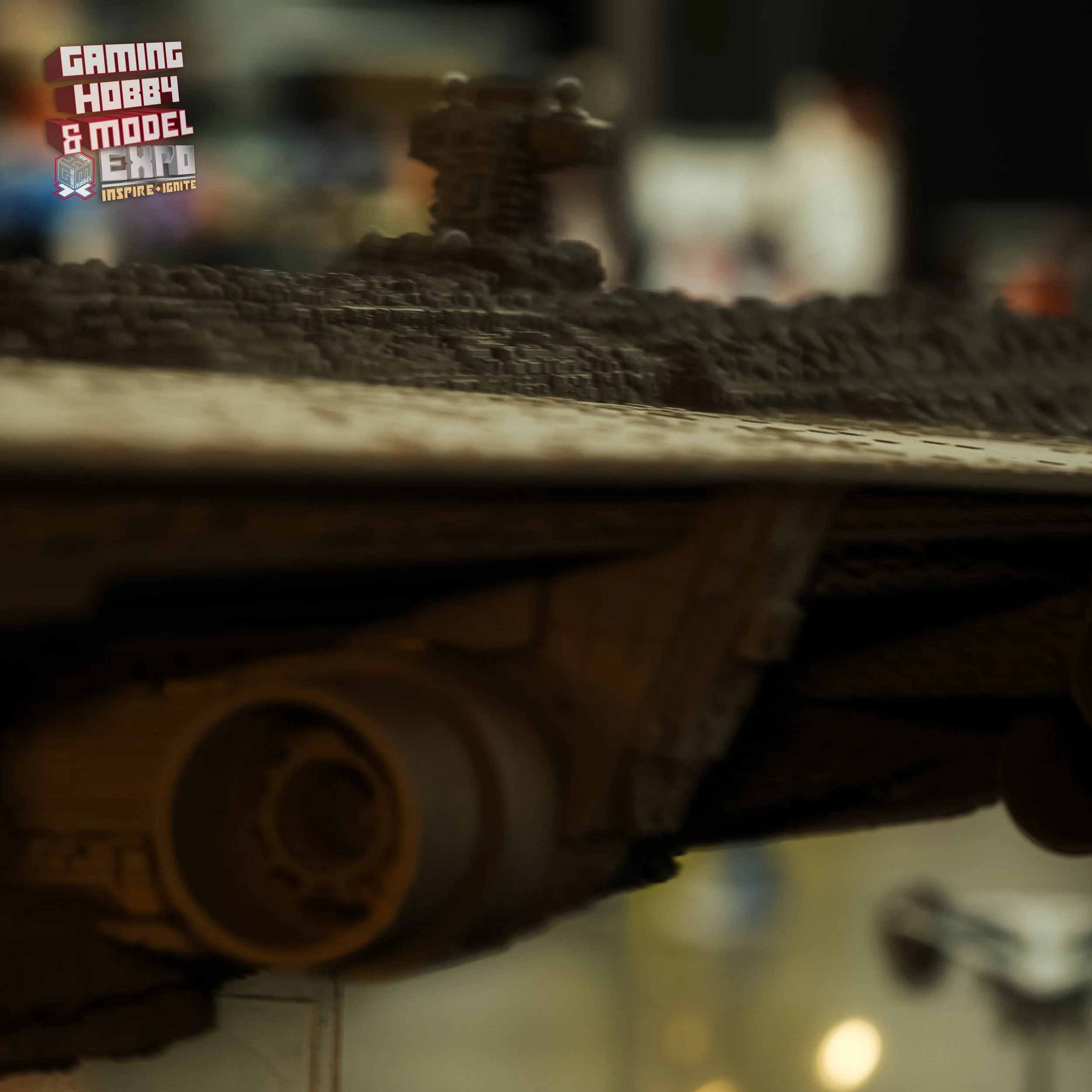 Close-up view of a detailed model spaceship or aircraft, with intricate surface textures and components, displayed on a flat surface. The background features a blurred logo or sign displaying 'Gaming Hobby & Model Expo' in colorful text.