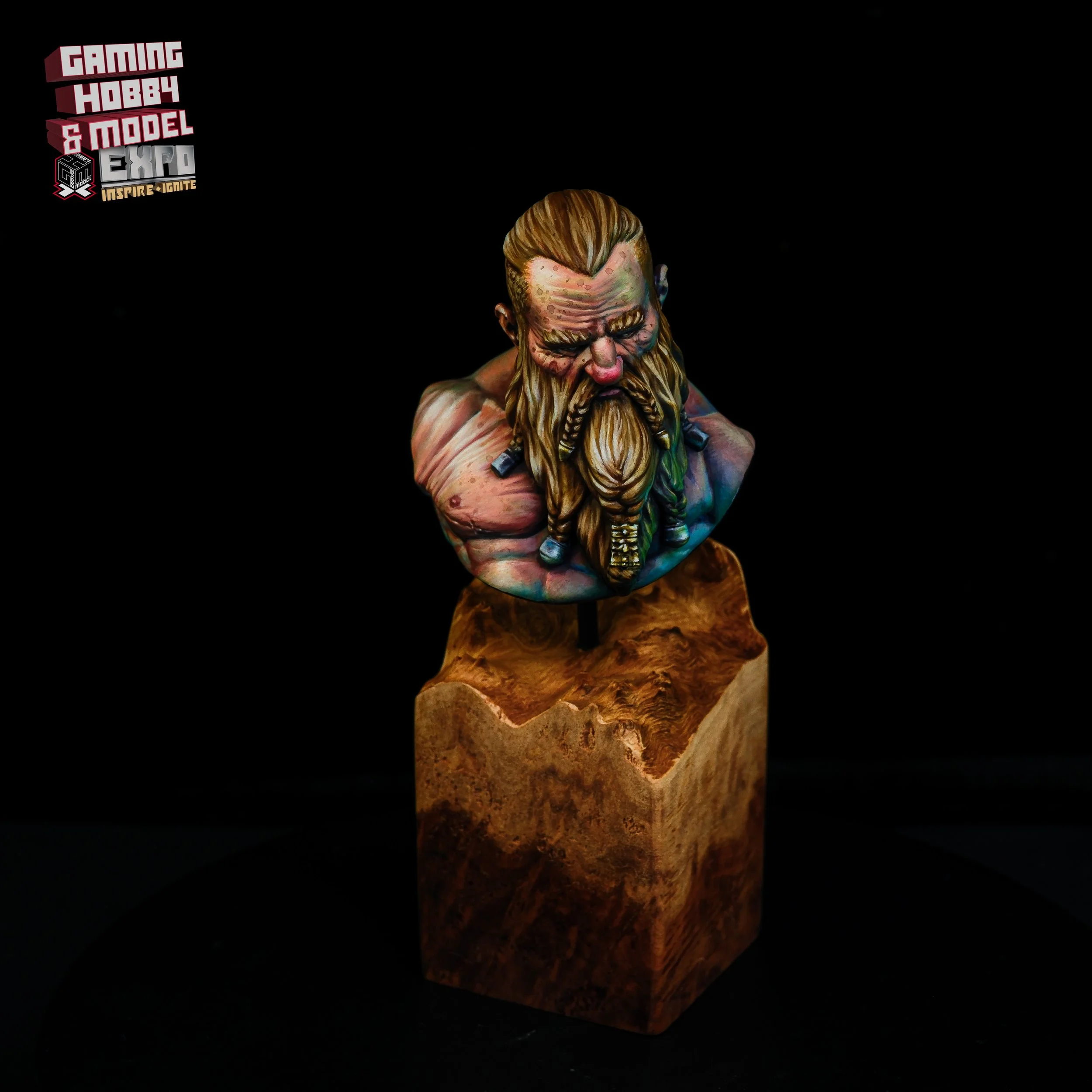A colorful bust sculpture of a bearded Viking warrior with long hair, mounted on a wooden stand, against a black background, at a gaming, hobby, and model expo.