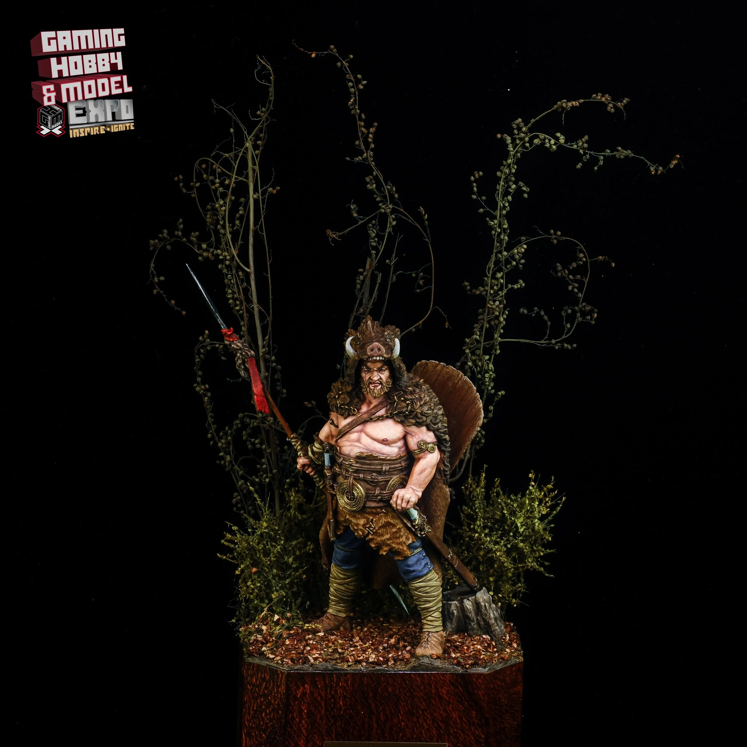 A detailed miniature figurine of a warrior with a pig head helmet, shirtless with armor, holding a spear, standing on a textured base with foliage and branches, at a hobby and model expo.