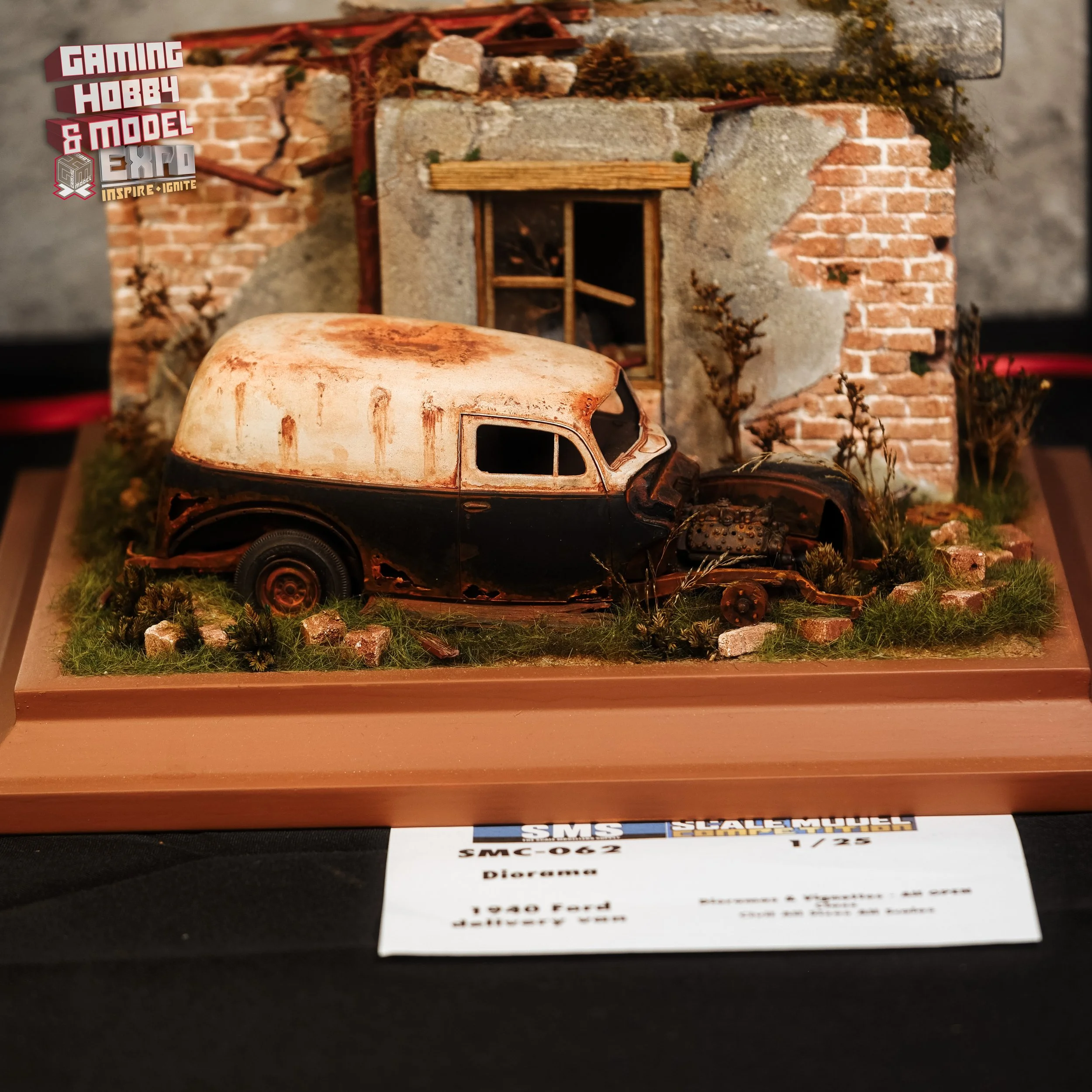 A detailed miniature diorama of an abandoned, rusted vintage 1940 Ford vehicle next to a weathered brick and stone building, surrounded by overgrown grass and small rocks.