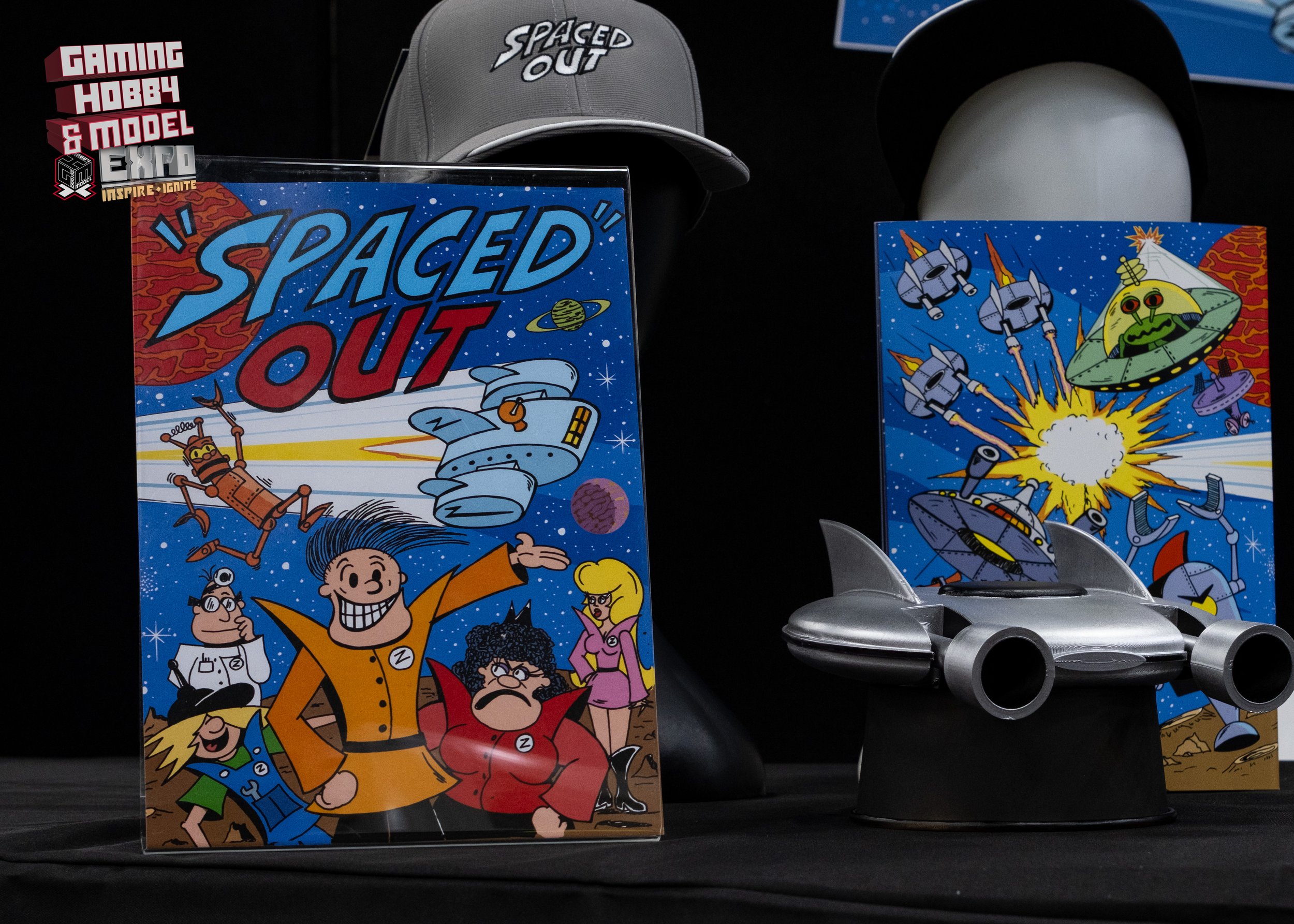 Space-themed promotional items including a framed comic book titled 'Spaced Out' featuring characters and space ships, a gray hat with 'Spaced Out' text, a sci-fi poster with explosions, robotic and alien spacecraft, and a small silver toy spaceship,