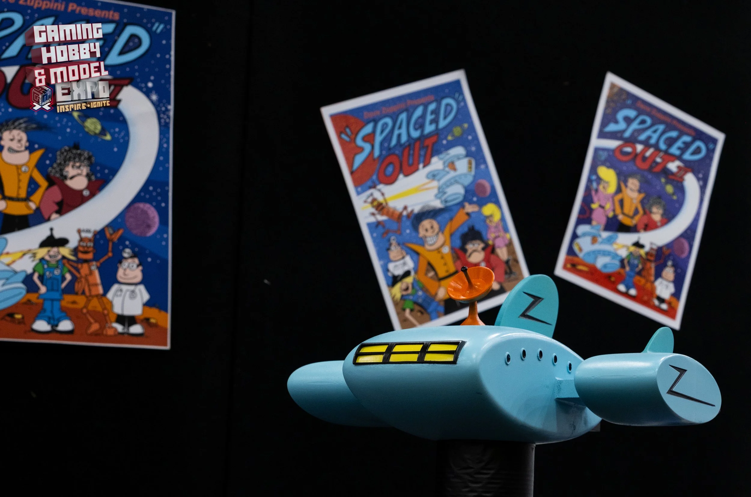 A blue spaceship-shaped toy with the letter 'Z' on its fins, situated in front of posters for 'Spaced Out' and 'Spaced Out' comics, printed with space-themed illustrations and characters, displayed against a dark background.