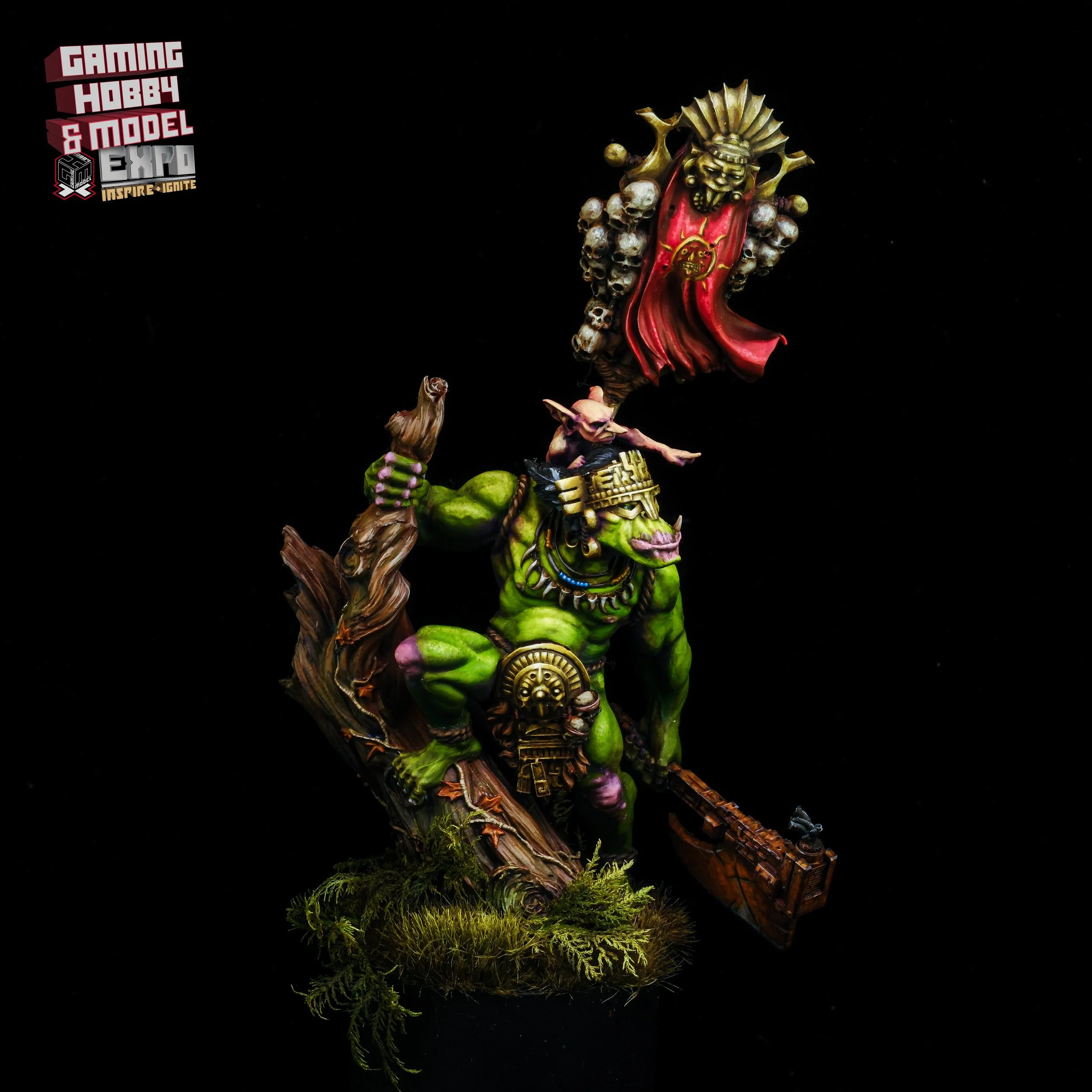A detailed miniature model of a green orc warrior with tribal jewelry, armor, and weapons, positioned on a base with moss and wood debris, with a dark background and a logo in the top left corner.