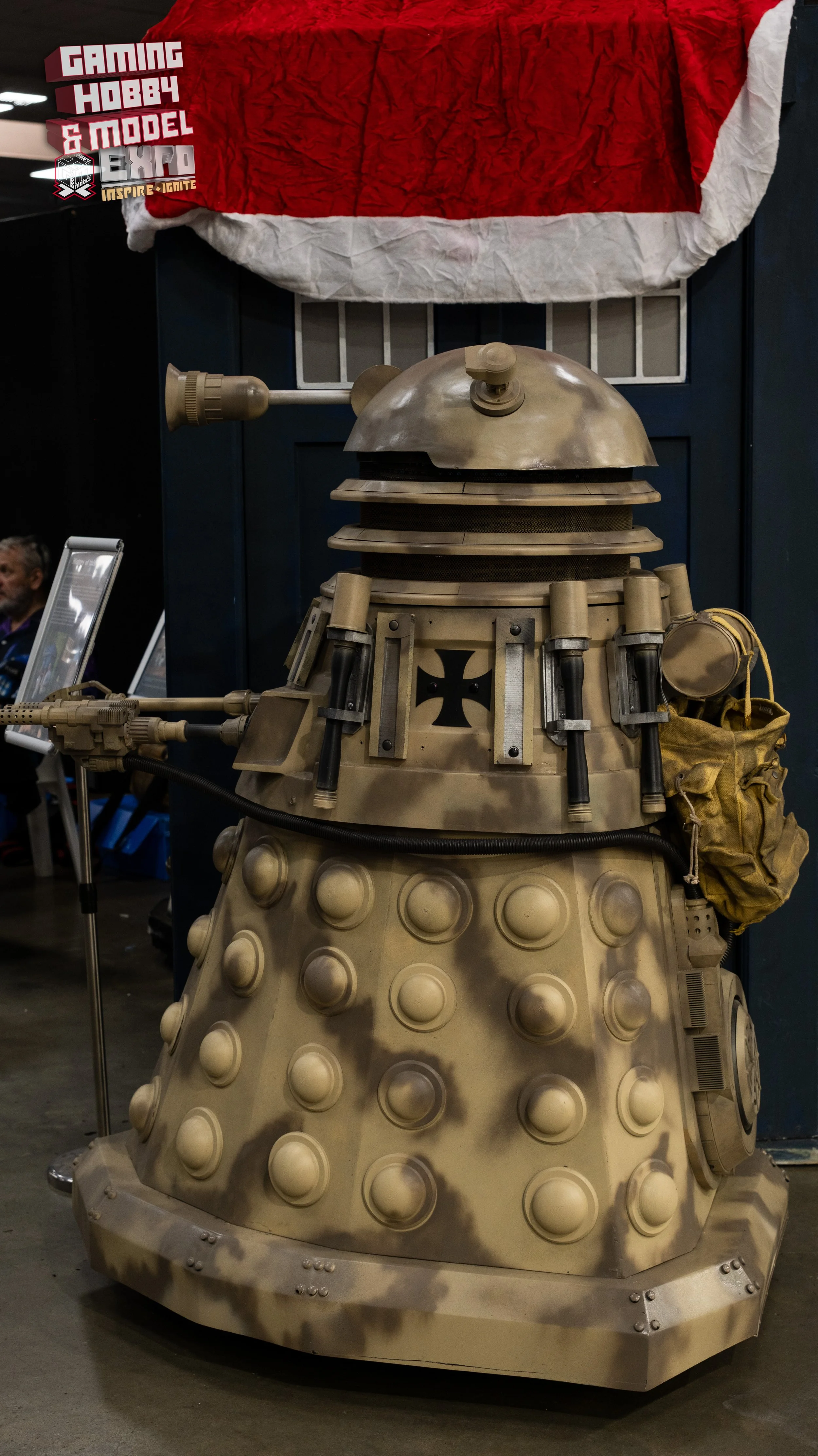 Life-sized replica of a Dalek from Doctor Who, painted in desert camouflage colors, with a raised arm and a bag hanging on the side. There is a red and white Santa hat draped above it, and a sign for a gaming hobby and model expo in the background.