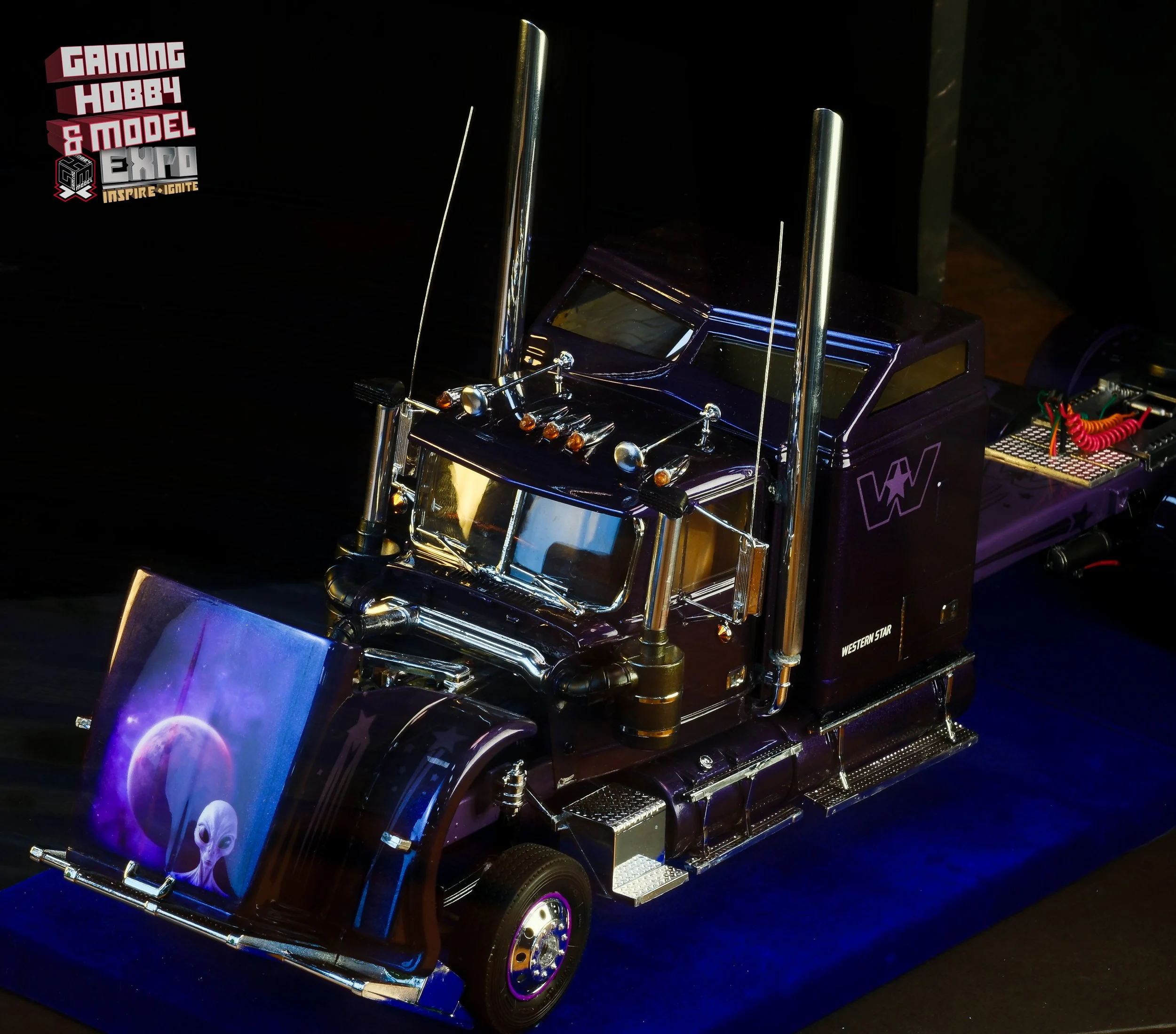 A detailed model of a semi-truck with a purple and black color scheme, featuring chrome accents, antennas, and a painted front bumper with an alien and moon landscape artwork, displayed at a gaming hobby and model expo.